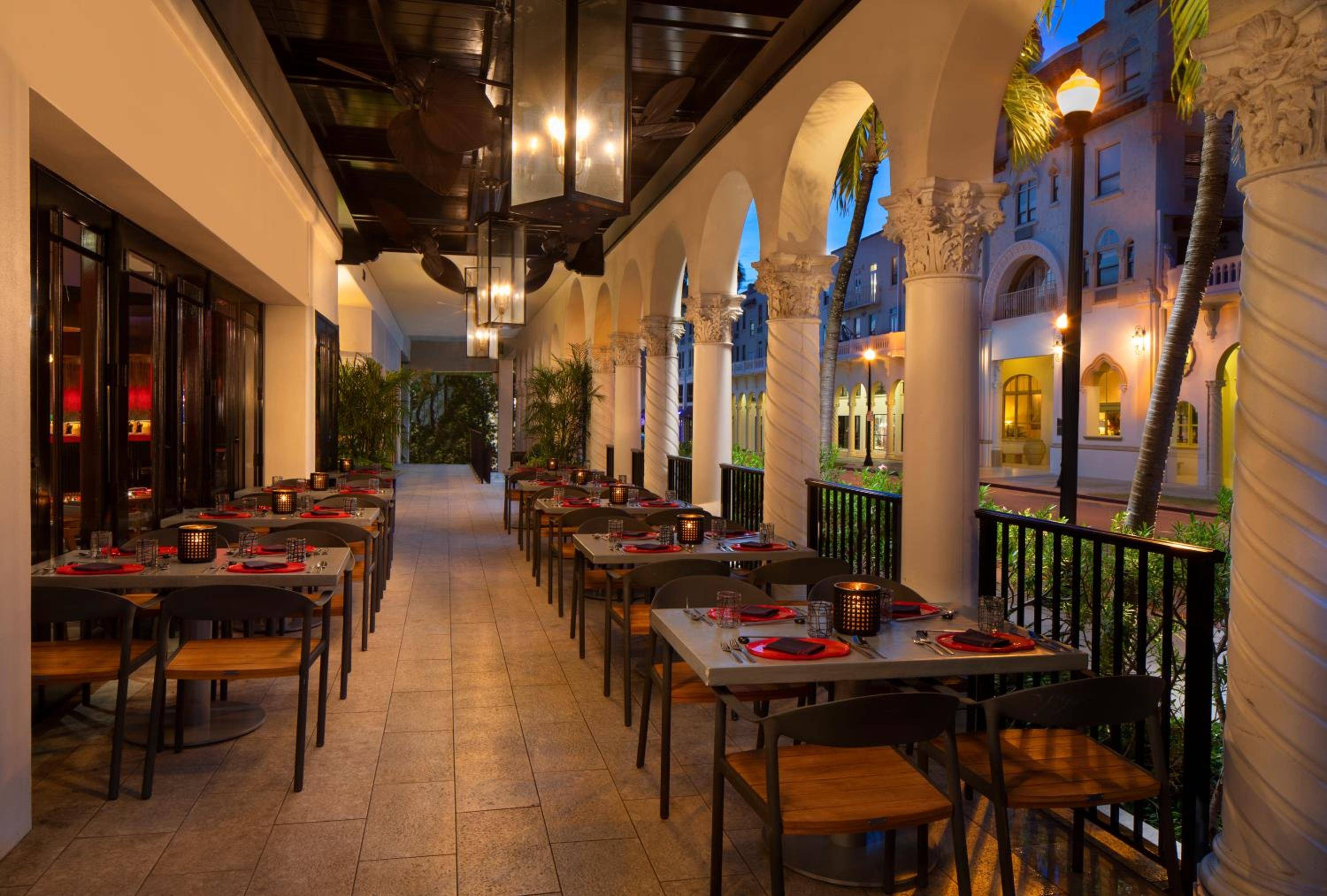 Flickering lanterns cast warm shadows across the colonnade terrace, where red placemats and dark wood tables create intimate dining beneath soaring arches overlooking the courtyard.