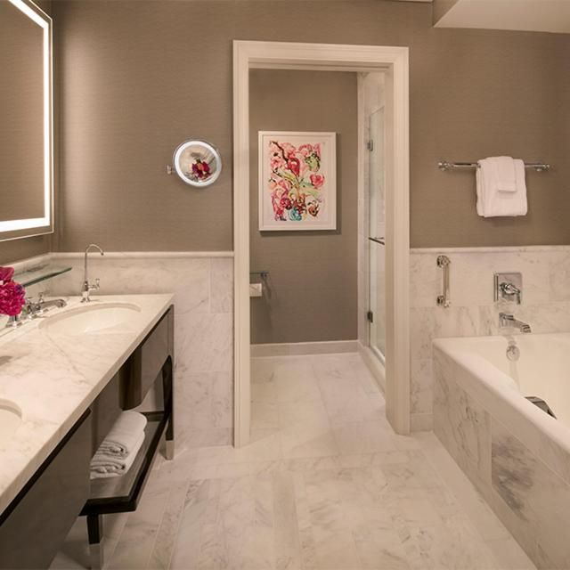 Warm taupe walls and pristine Carrara marble create a spa-like sanctuary where floral artwork adds unexpected charm to this luxurious soaking retreat.