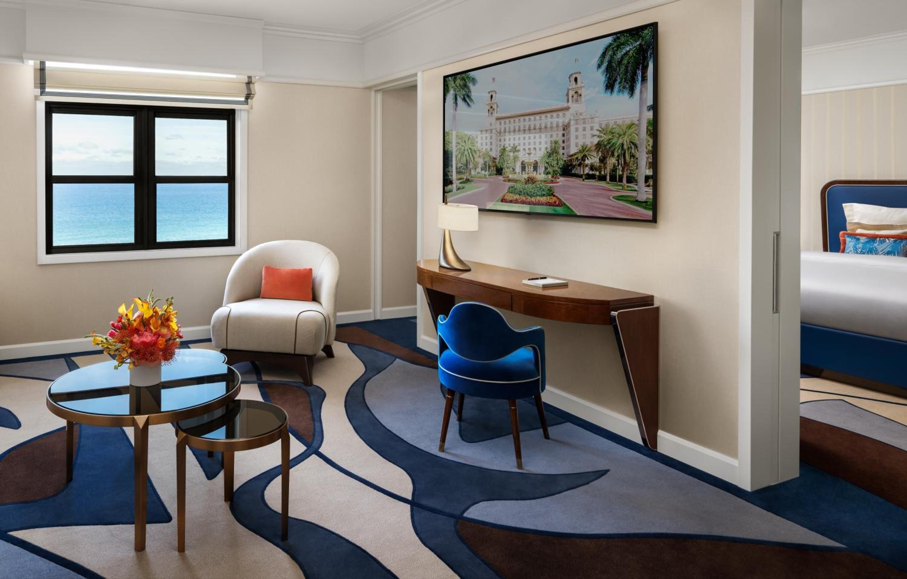 Ocean views frame this sophisticated suite where navy geometric carpeting anchors walnut furnishings and a coral accent pillow, while palm-fringed artwork celebrates the resort's storied elegance.