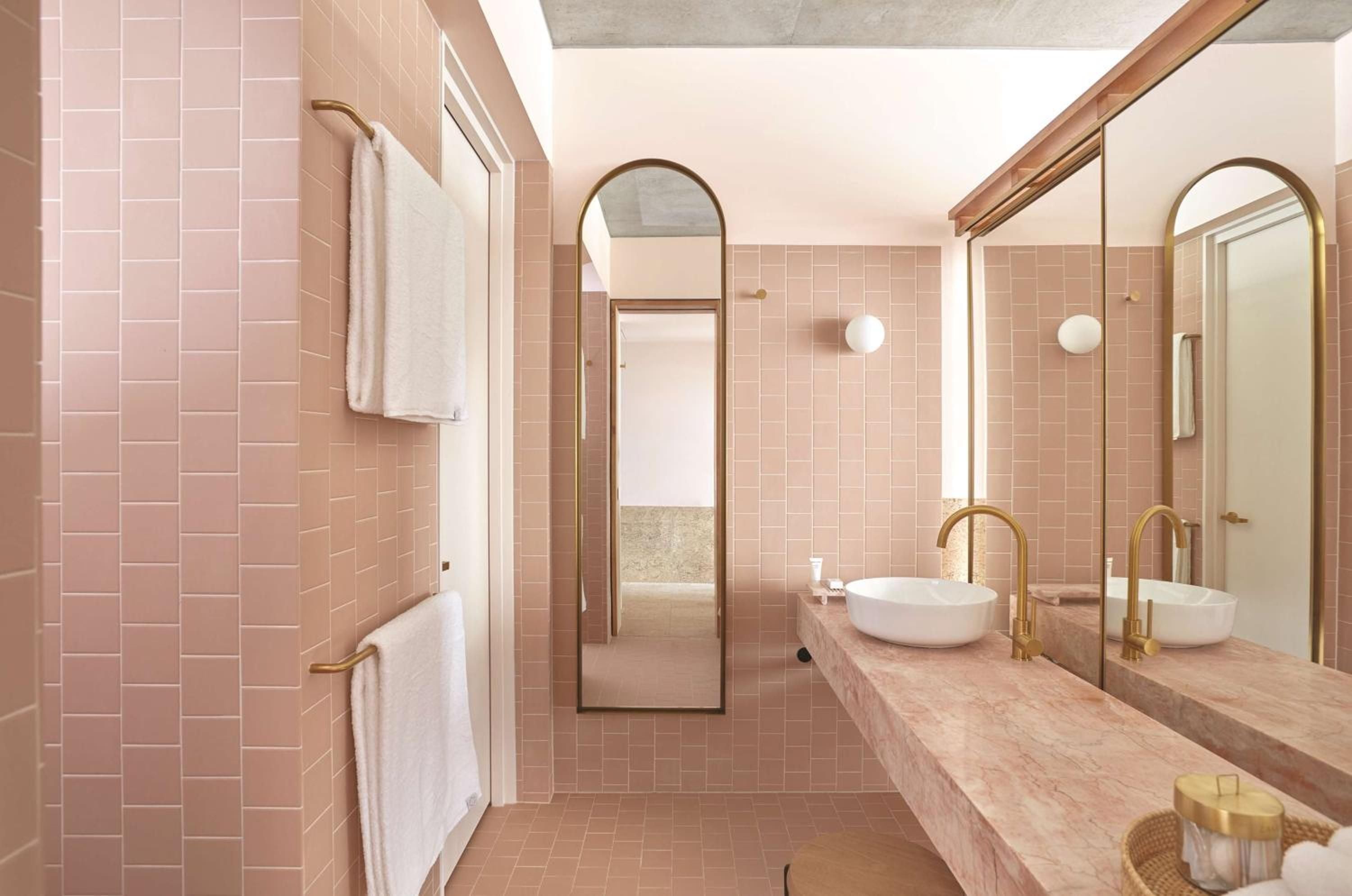 Blush-pink tiles and brass fixtures create an intimate sanctuary where vessel sinks rest on natural stone, reflecting warm light through arched mirrors.