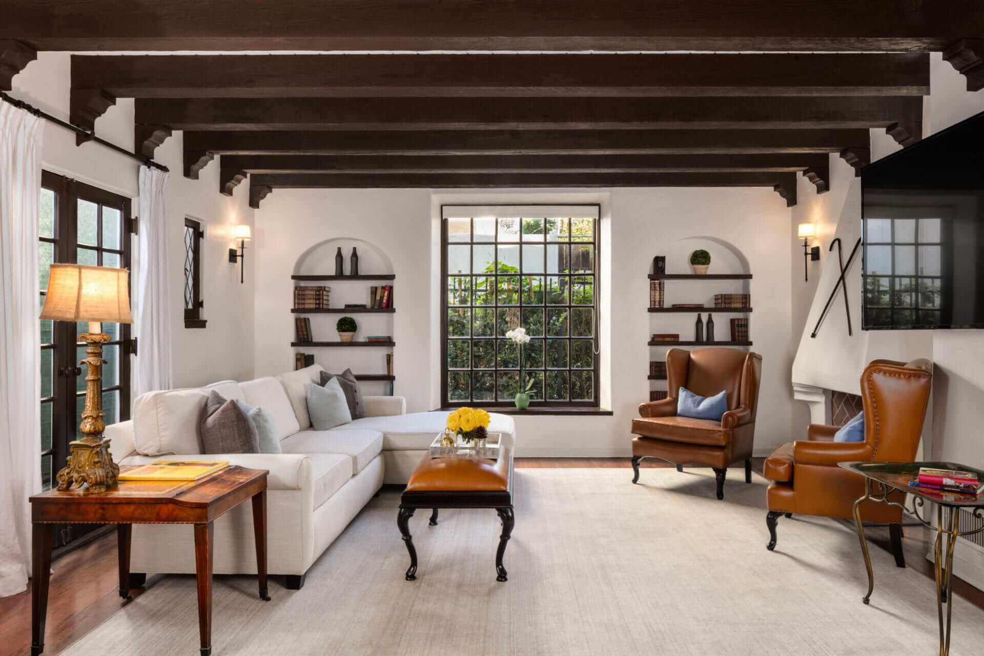 Exposed wooden beams frame this sophisticated living space where cognac leather wingbacks and built-in shelving create an inviting sanctuary flooded with natural light.