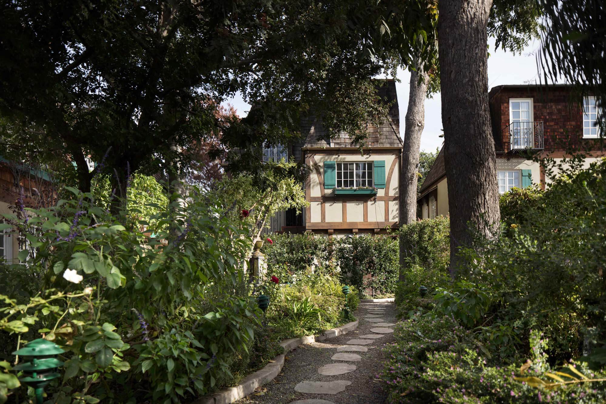 A winding stone path beckons through lush cottage gardens, where timber-framed buildings with mint-green shutters peek through dappled sunlight and mature oak canopies.