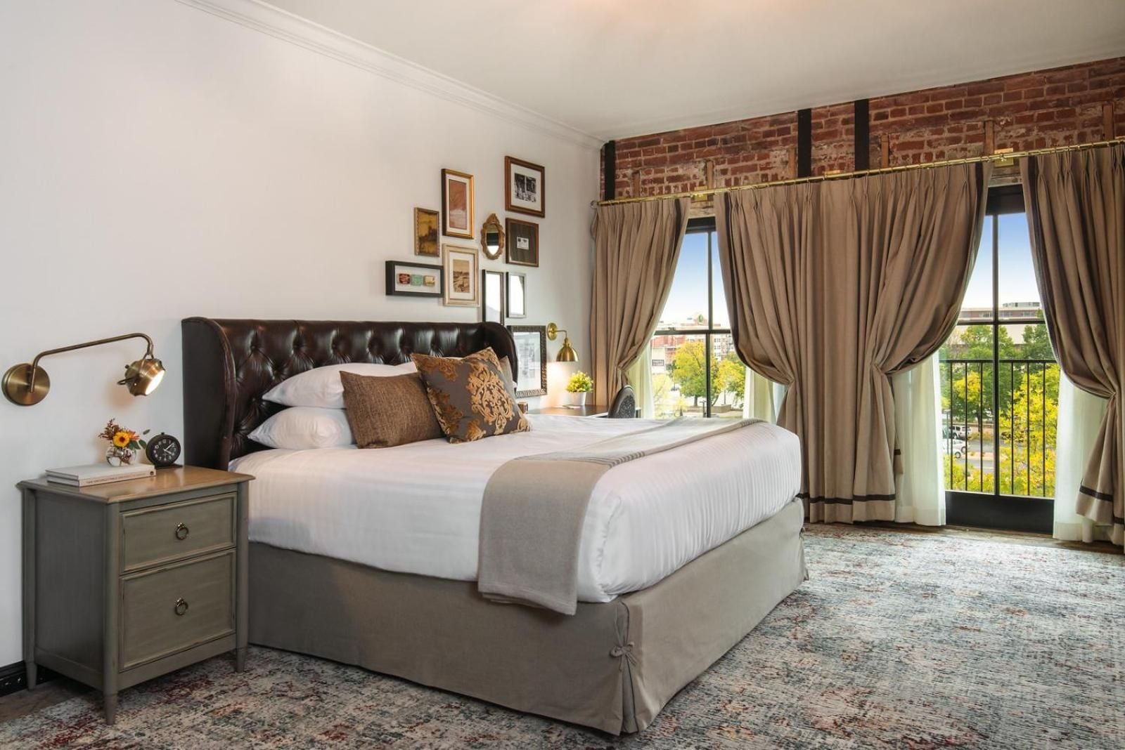 Exposed brick walls and brass sconces create intimate warmth in this thoughtfully appointed guest room, where French doors frame tree-lined views beyond vintage-inspired furnishings.