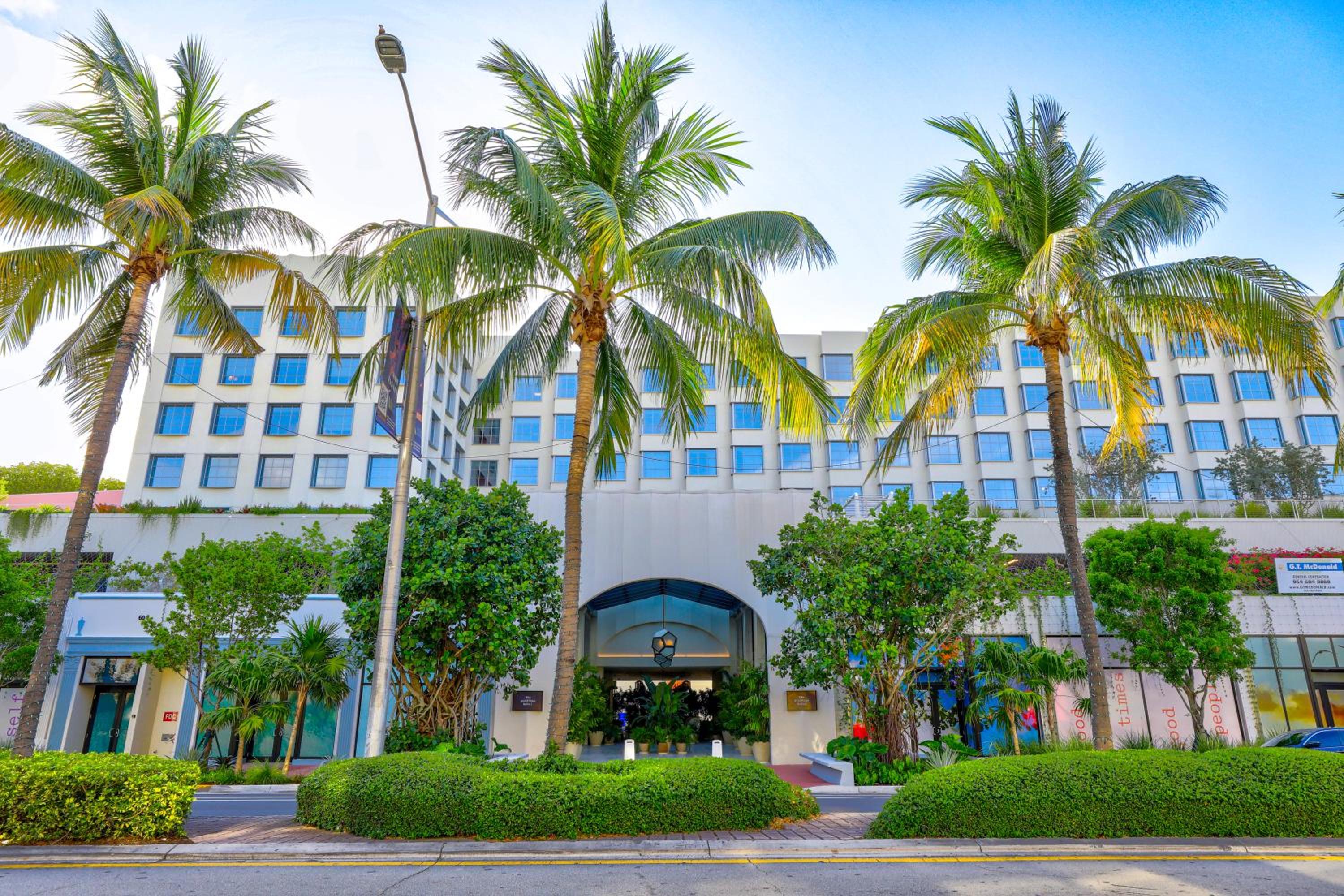 Swaying coconut palms frame the hotel's crisp white facade, where manicured hedgerows lead to an inviting arched entrance beneath Miami's endless blue sky.