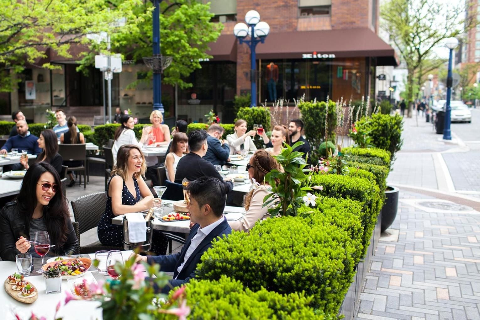 Guests savor cocktails and refined cuisine along the bustling sidewalk patio, where manicured hedges create intimate dining nooks amid the urban energy.