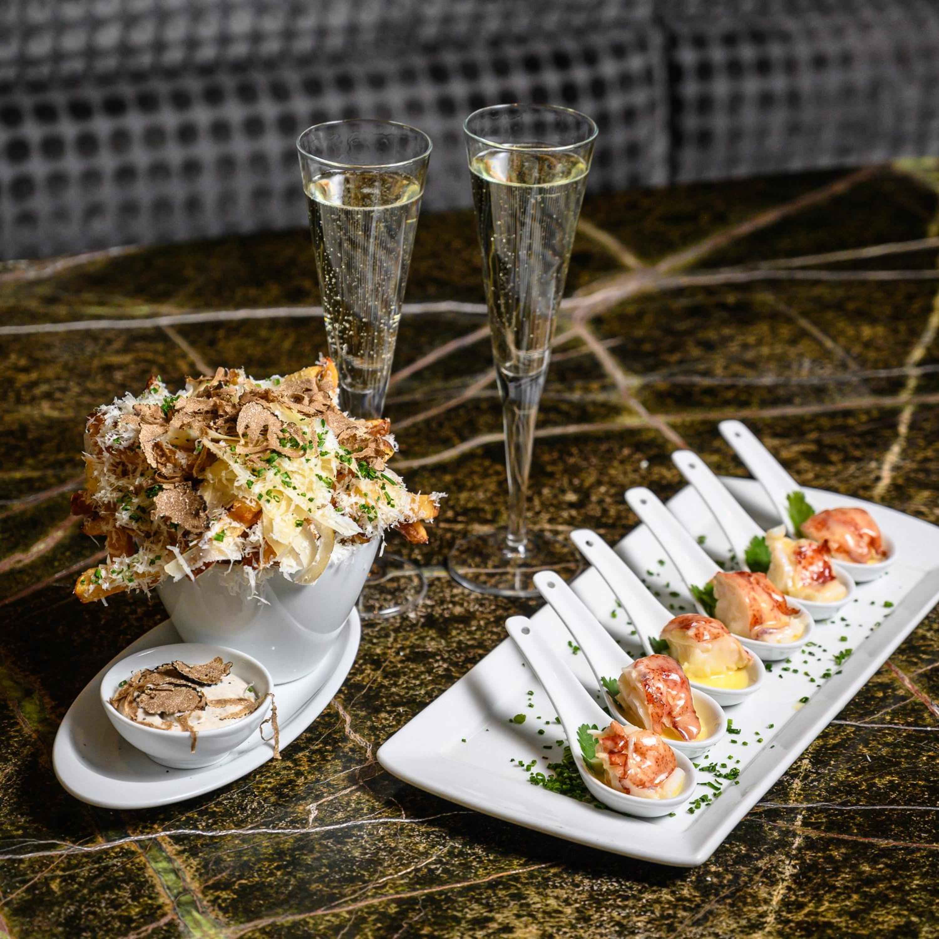 Champagne flutes catch golden light beside truffle pasta and elegant canapés, where culinary artistry meets sparkling celebration on gleaming marble surfaces.