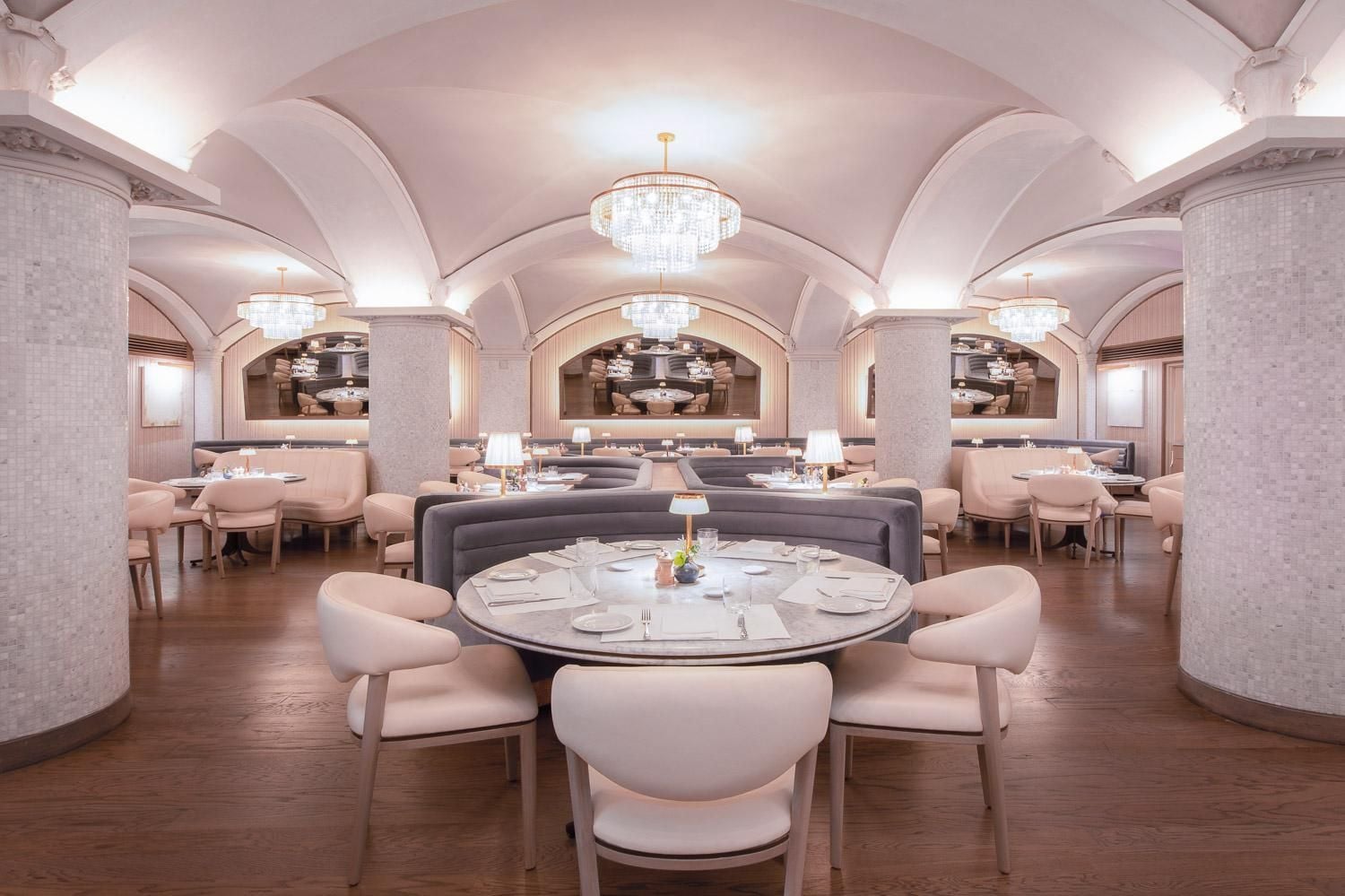 Vaulted ceilings and crystal chandeliers create cathedral-like grandeur in the dining room, where blush velvet chairs gather around pristine white tables on gleaming hardwood floors.