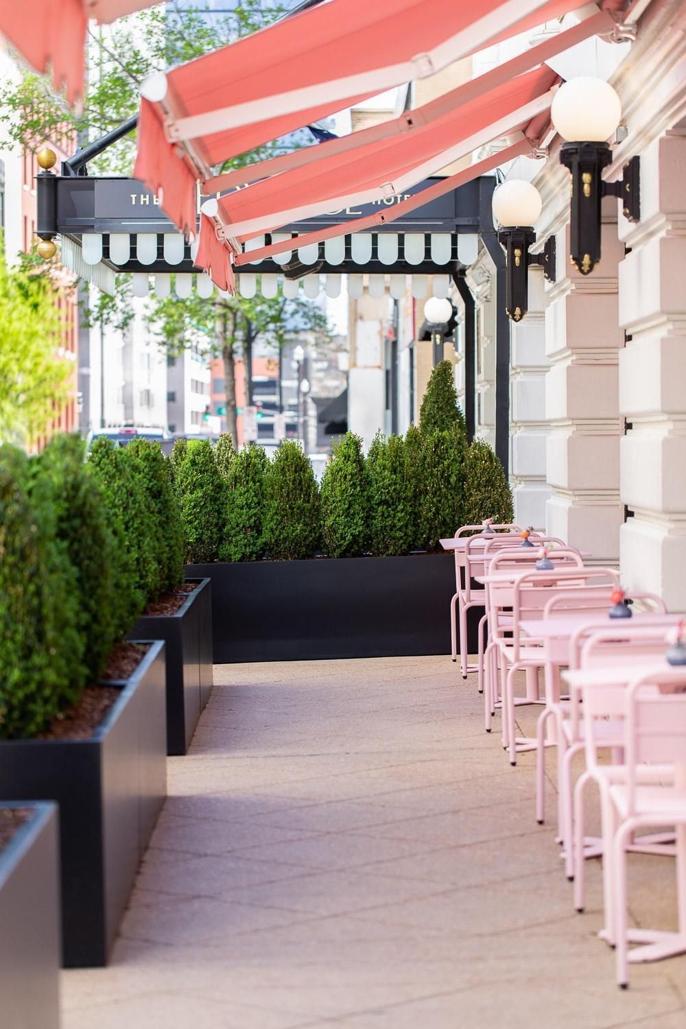 Coral-striped awnings shelter pink metal chairs along the sidewalk terrace, where manicured boxwood hedges create intimate dining nooks beneath vintage-style globe lanterns.