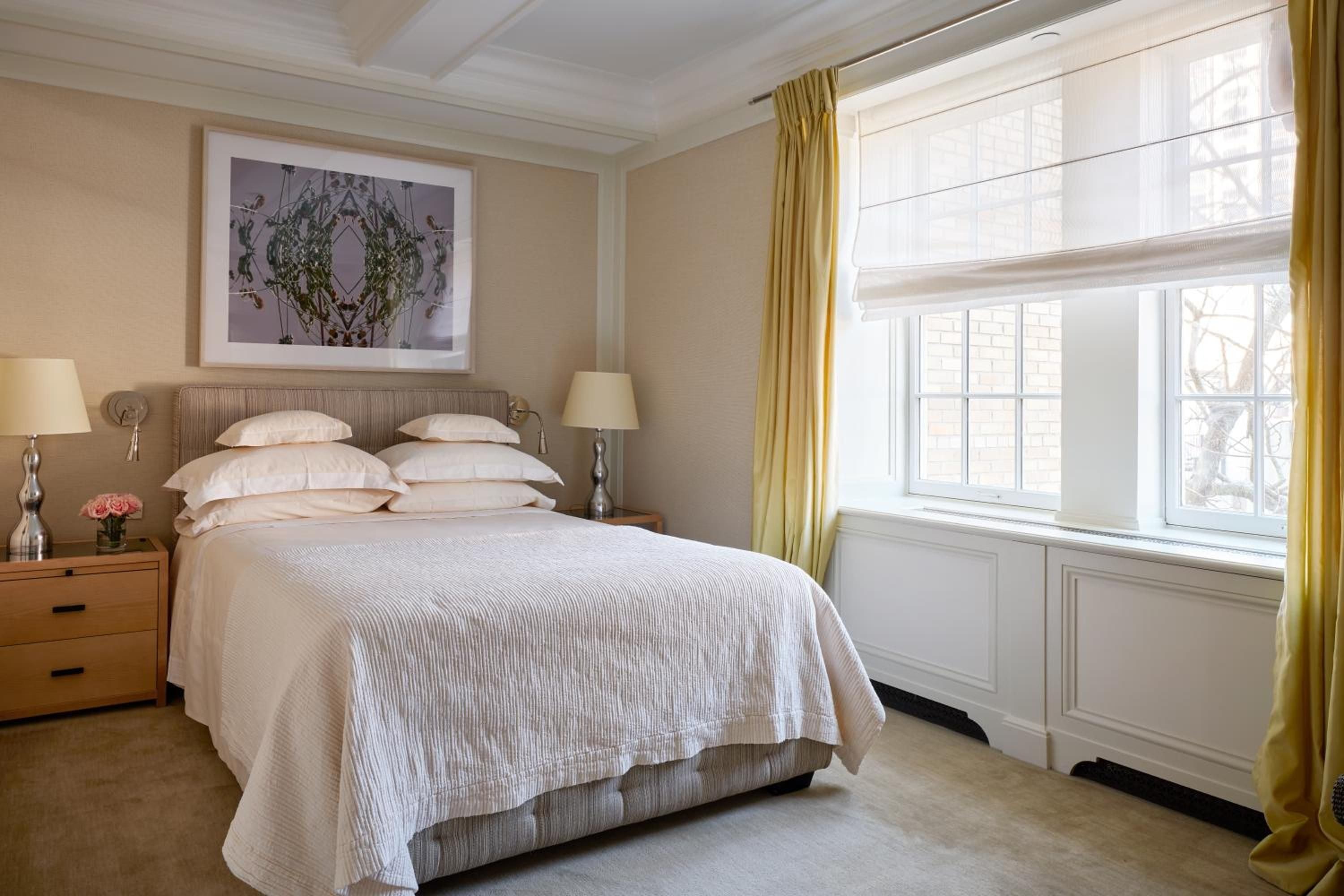 Soft morning light filters through crisp white Roman shades, illuminating neutral linens and warm wood furnishings in this serene guest room sanctuary.