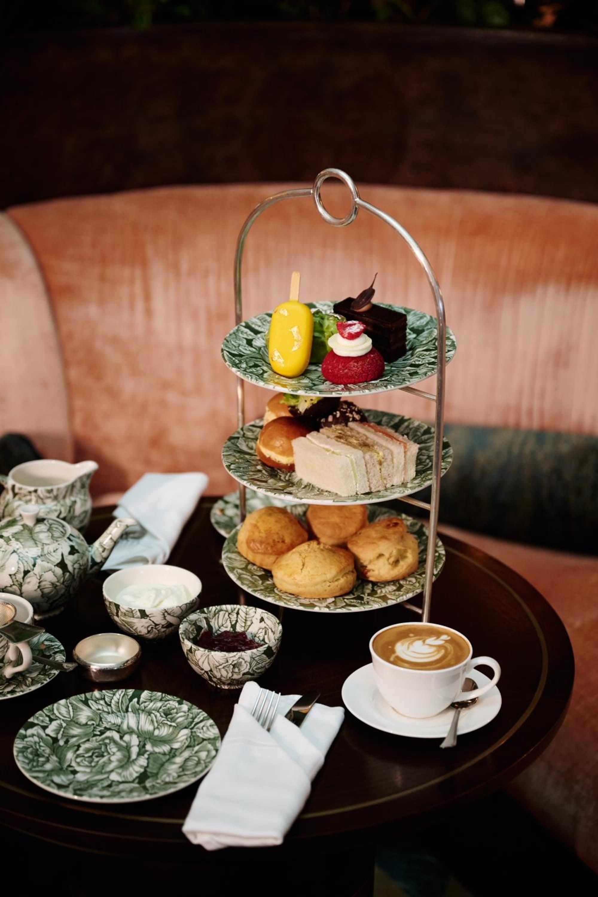 Elegant afternoon tea service unfolds on botanical-patterned china, where delicate petit fours and golden scones create an intimate ritual against plush coral banquettes.