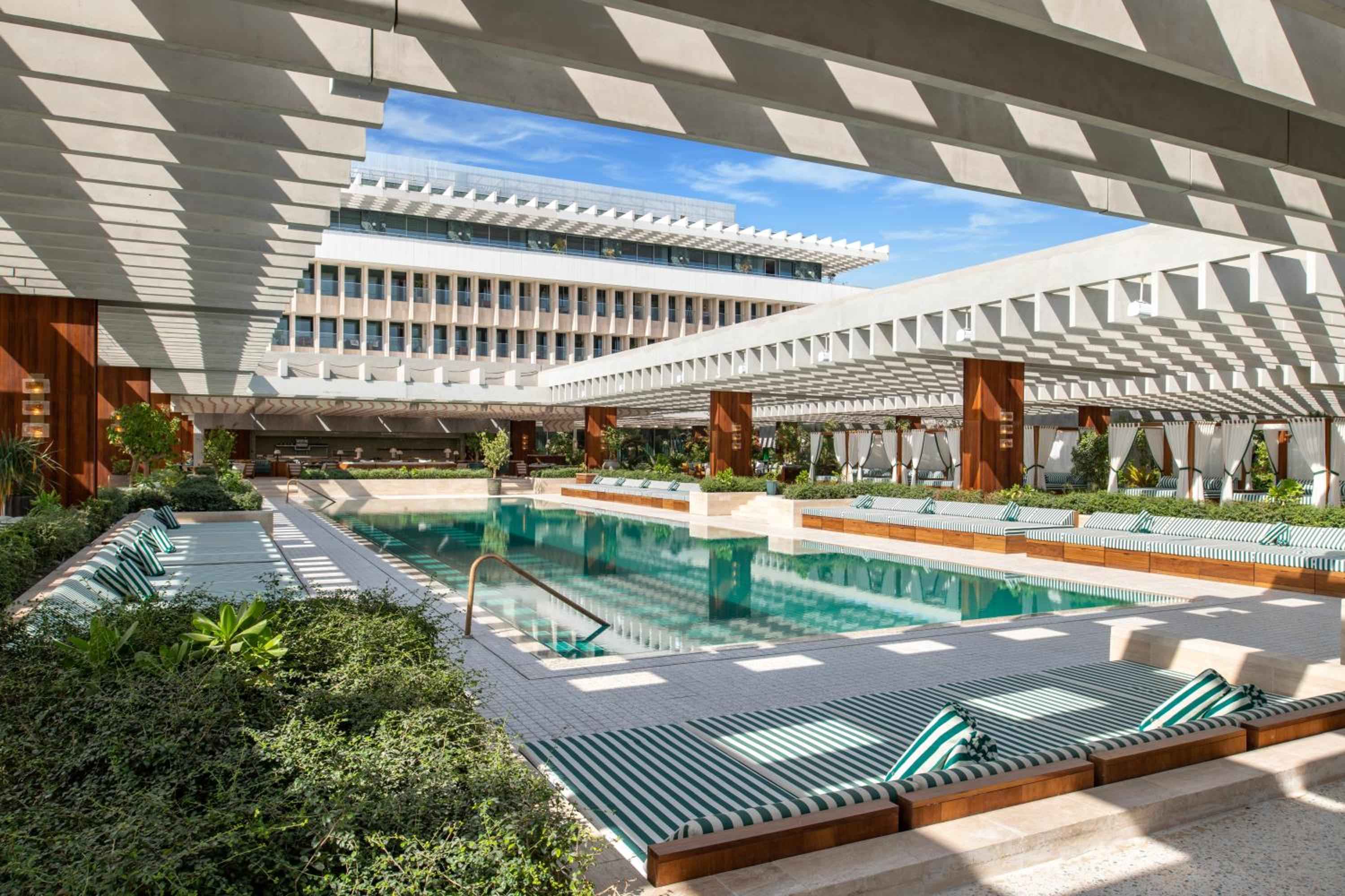 Geometric shadows dance across striped loungers at the rooftop pool, where white latticed pergolas frame turquoise waters against Doha's gleaming skyline.