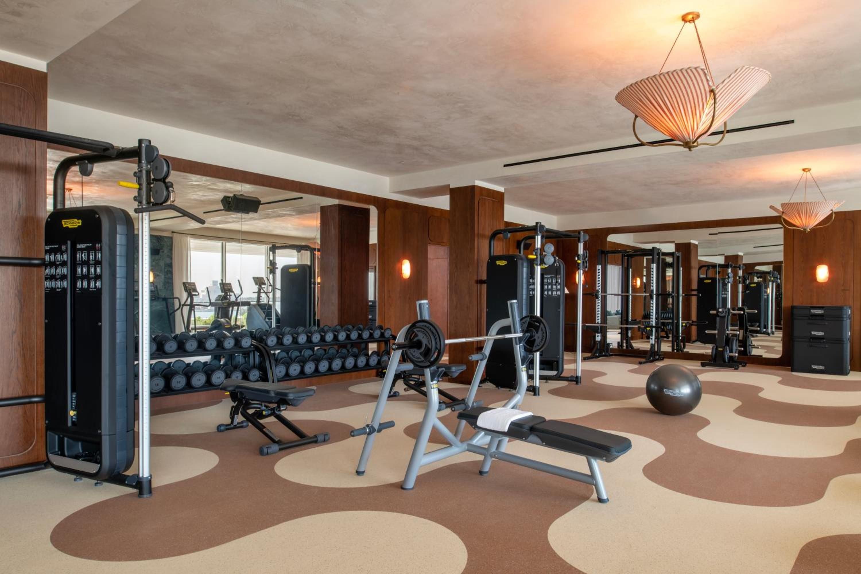 Sculptural pendant lights cast warm glows over polished equipment in this striking fitness center, where geometric flooring patterns in earthy tones create an unexpectedly sophisticated workout environment.