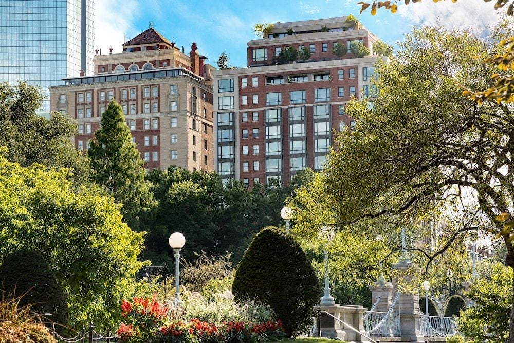 Historic brick facades rise gracefully above Boston Public Garden's manicured topiaries and seasonal blooms, where city sophistication meets pastoral tranquility in the heart of Back Bay.