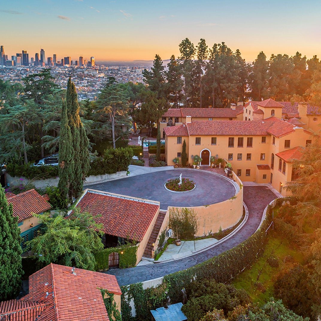 Golden hour light bathes the estate's distinctive circular courtyard and red-tiled roofs, while downtown LA's glittering skyline stretches beyond the lush hillside gardens.