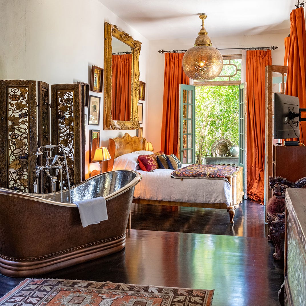 Moroccan-inspired luxury meets Hollywood glamour in this sumptuous suite, where vibrant orange silk curtains frame garden views and a copper bathtub anchors rich wood floors.