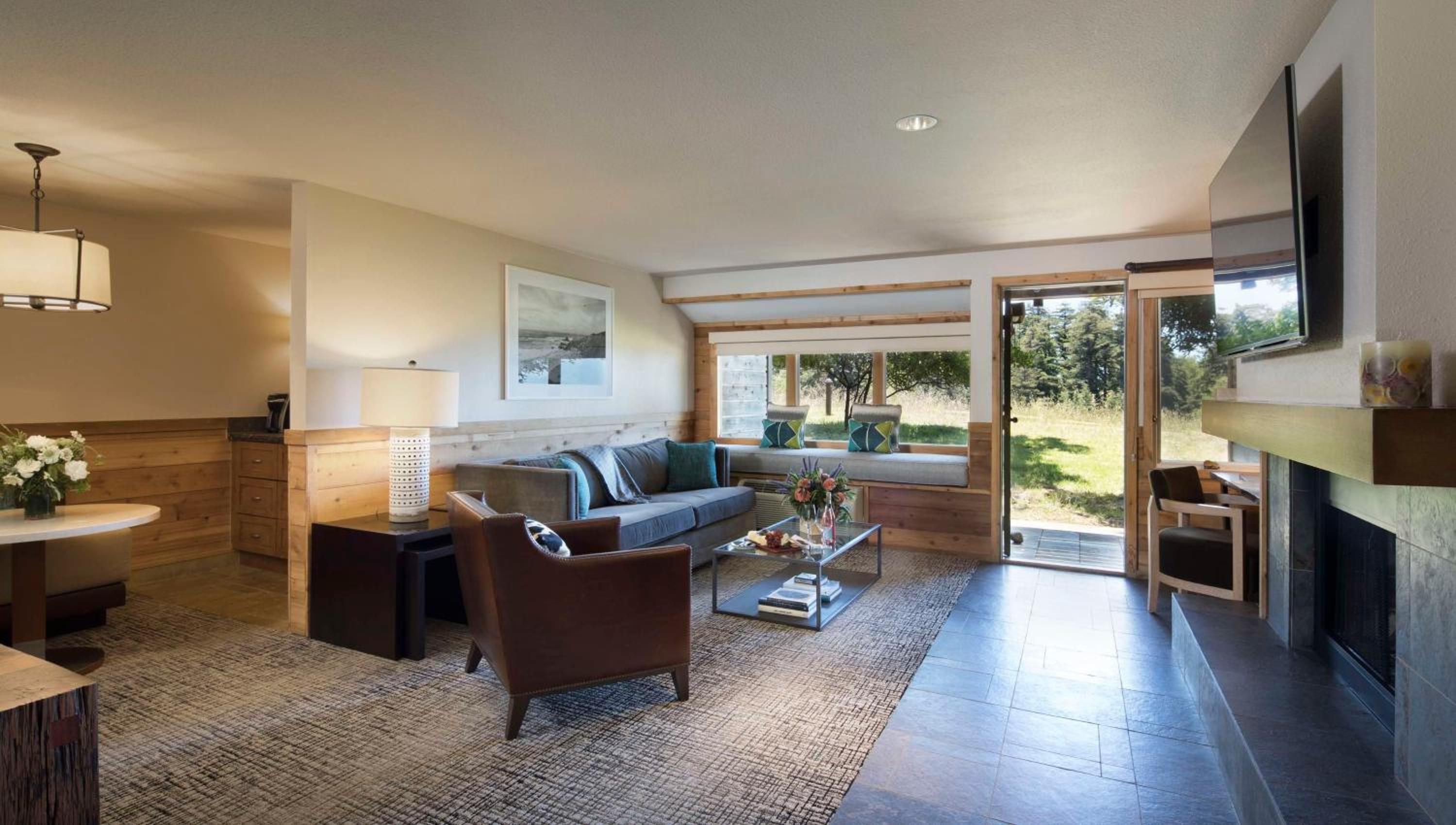 Warm wood tones and natural stone create an inviting retreat, where floor-to-ceiling windows frame Big Sur's wild landscape beyond the comfortable seating area.