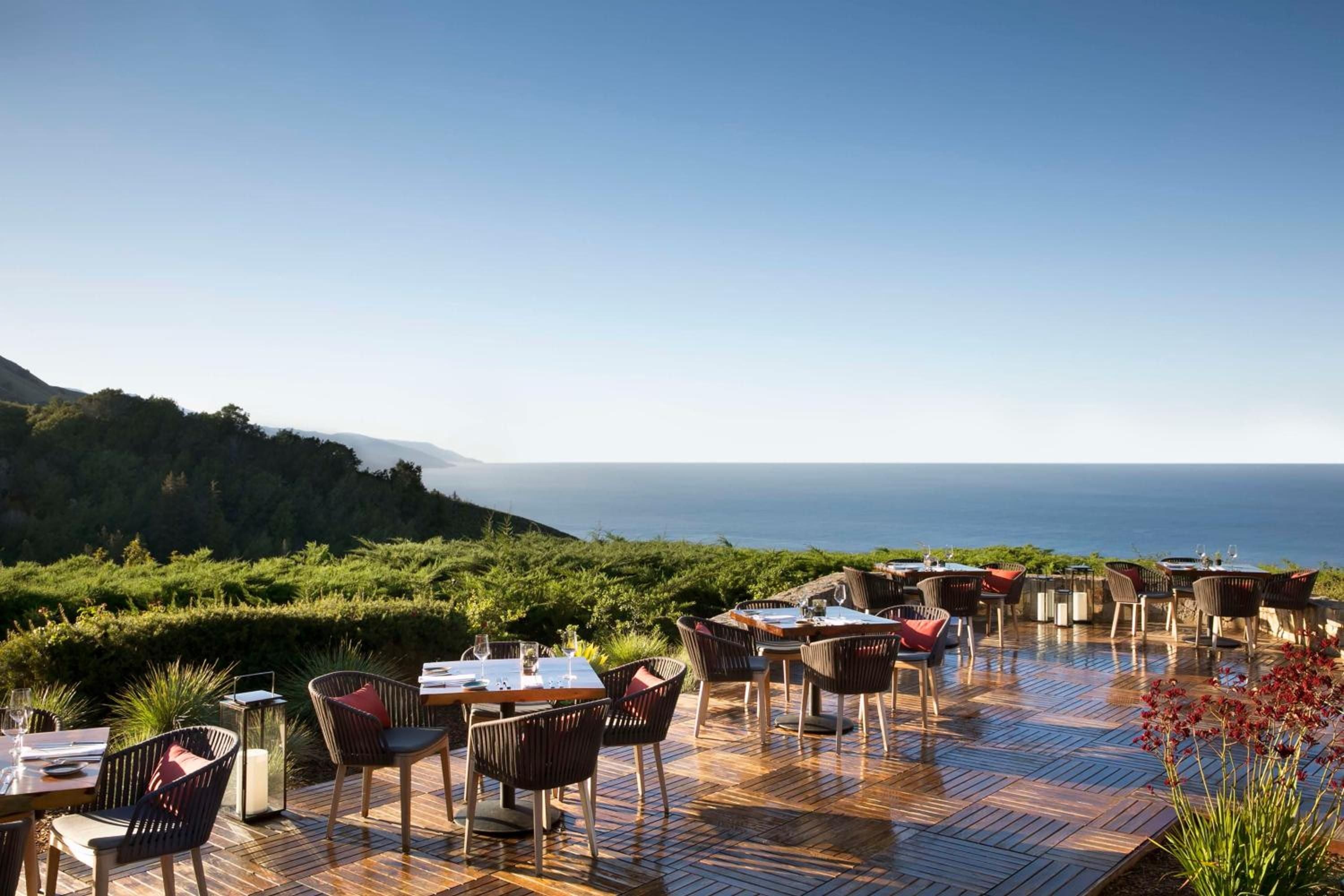 Weathered teak tables dot the oceanfront terrace where Big Sur's dramatic coastline unfolds beyond intimate dining spaces wrapped in native grasses and salt air.