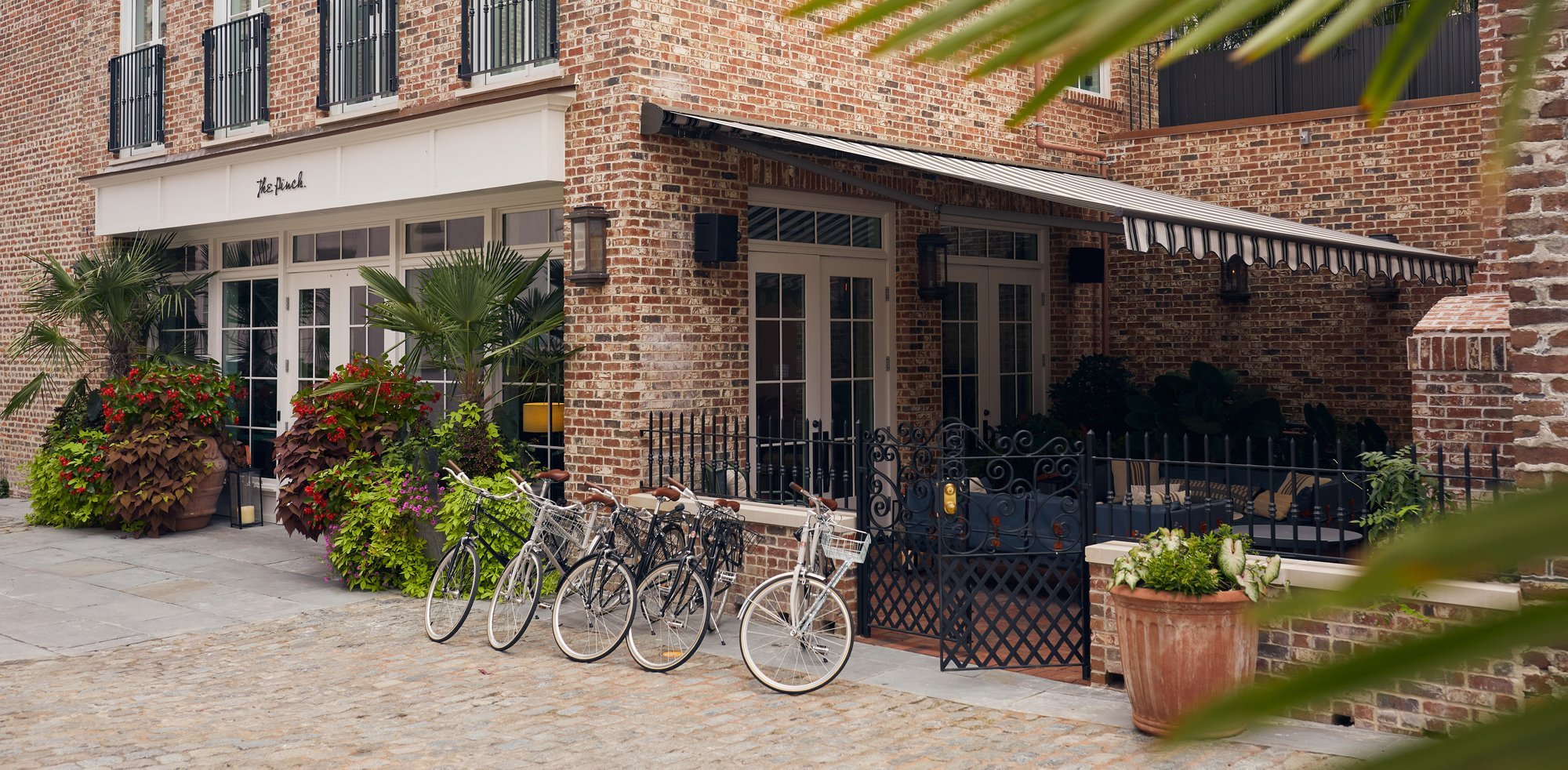 Vintage bicycles rest against weathered brick walls where Charleston's historic charm meets modern hospitality, creating an inviting streetscape of cobblestones and colorful plantings.