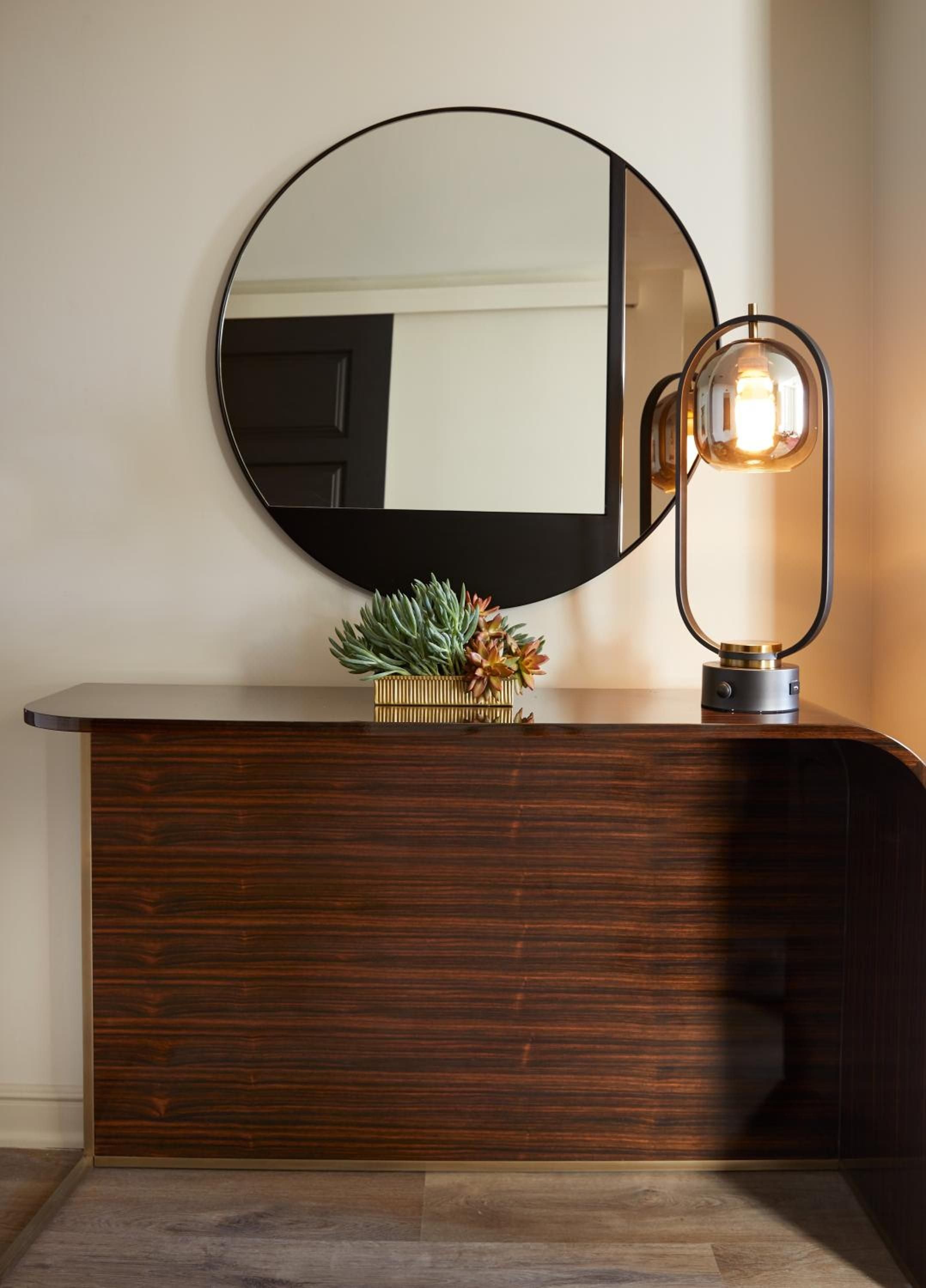 Warm amber light from a sculptural table lamp illuminates rich walnut wood grain and desert succulents, creating an intimate corner where modern luxury meets organic comfort.