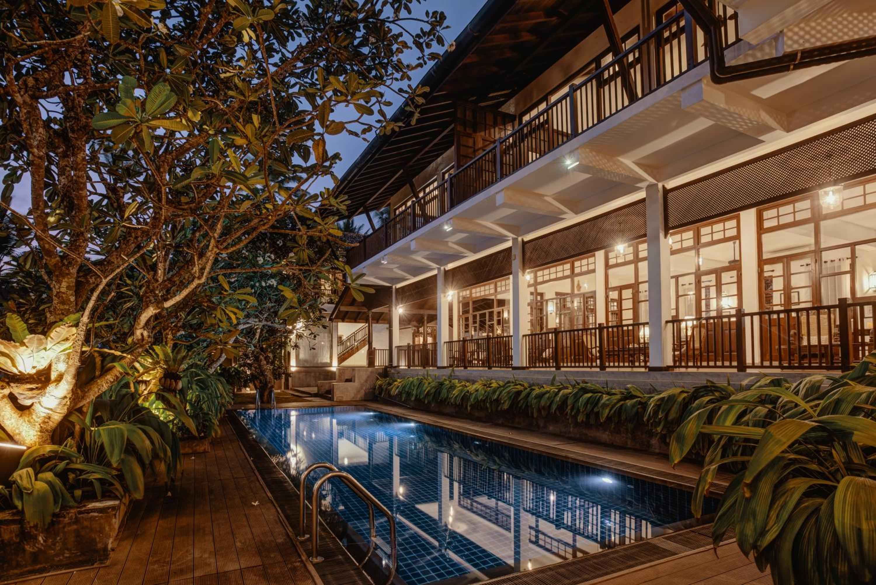 Colonial-style verandas embrace a jewel-toned pool where frangipani trees cast dancing shadows and warm light spills from timber-framed windows into the tropical evening.