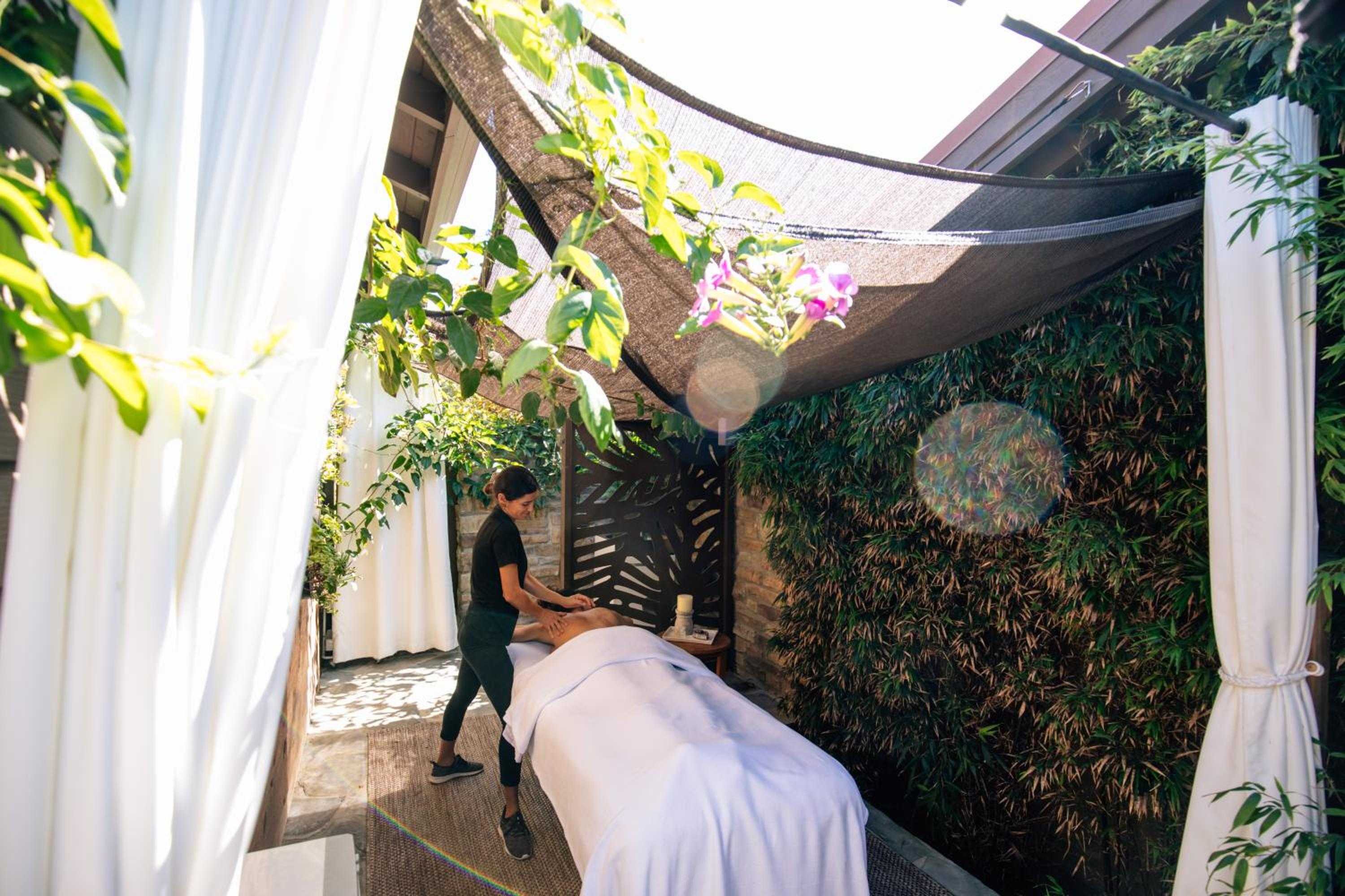 Dappled sunlight filters through shade sails as skilled hands work beneath swaying palms, creating an intimate outdoor sanctuary where massage meets garden tranquility.