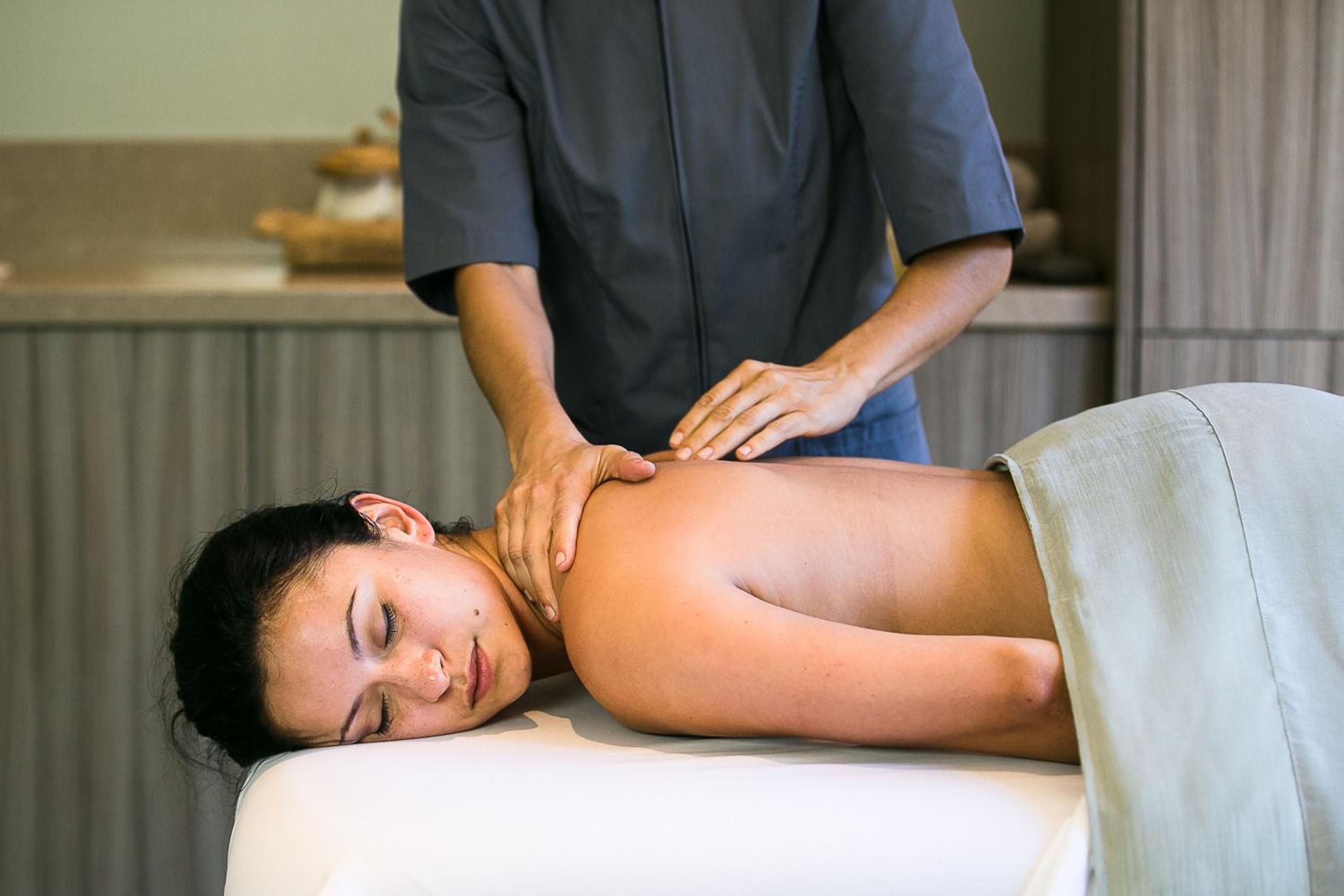 Expert hands work tension from shoulders and neck in the serene treatment rooms, where warm lighting and natural textures create an intimate wellness sanctuary.