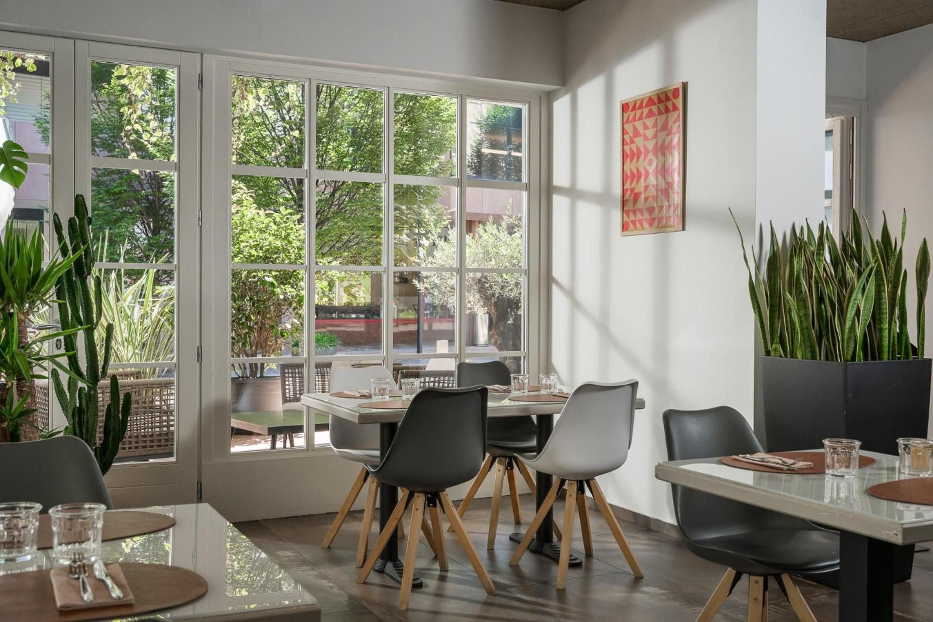 Modern dining chairs in muted tones gather around sleek tables, while tall snake plants frame floor-to-ceiling windows revealing the lush terrace beyond.