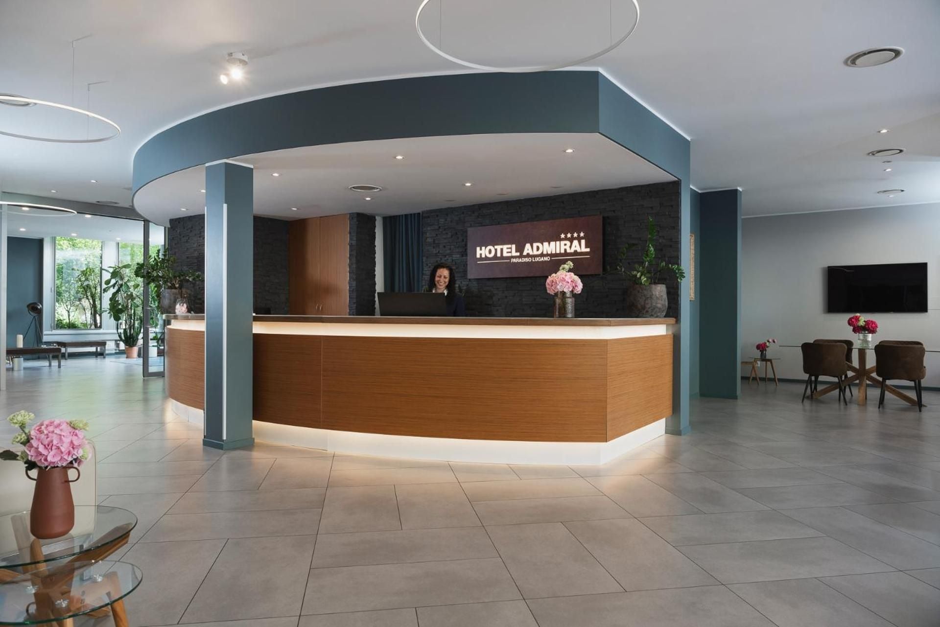 Hotel Admiral's curved reception desk anchors a sophisticated lobby where warm wood tones meet cool teal accents, pink peonies adding delicate color to the contemporary Swiss design.