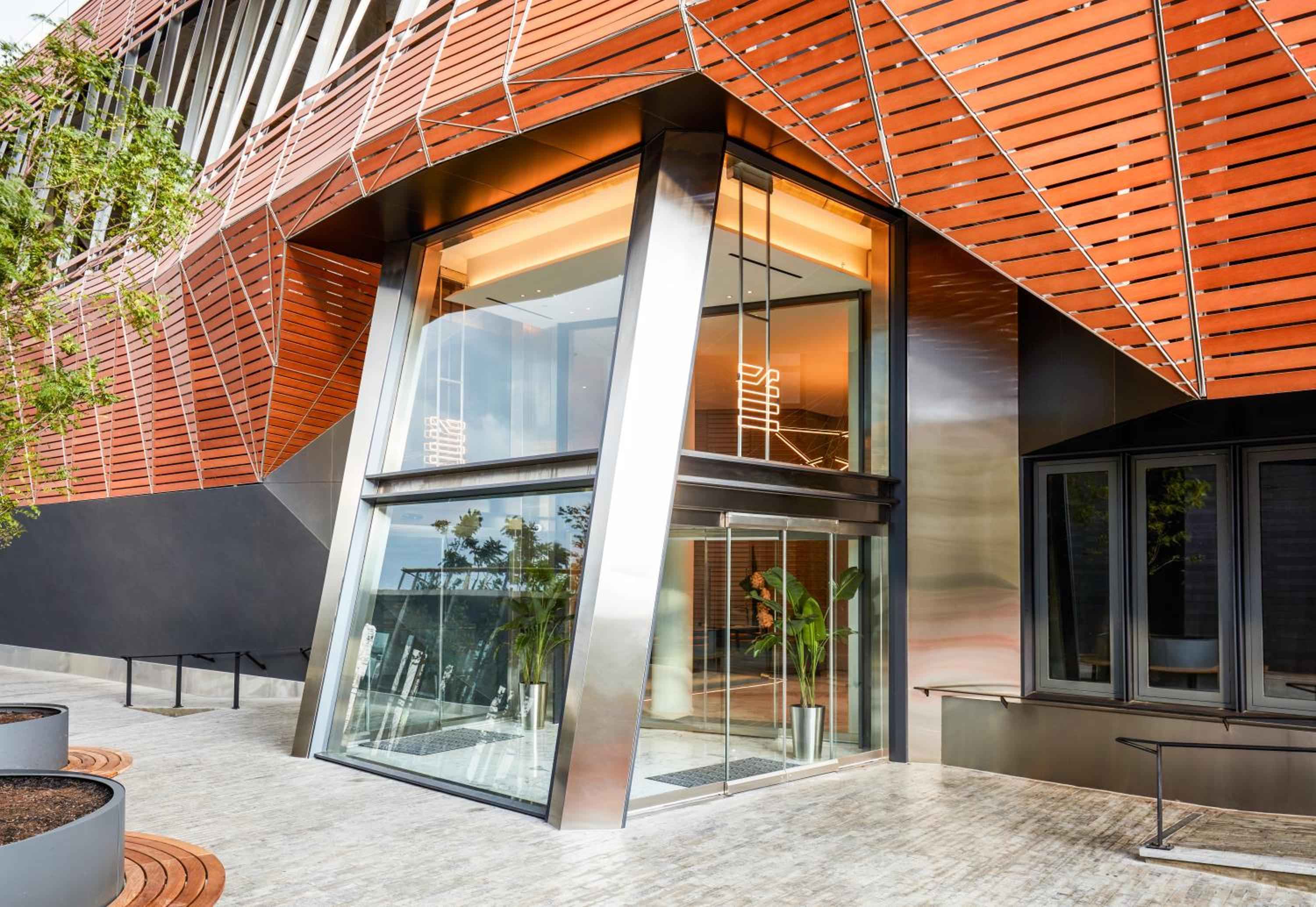 Warm wood slats cascade across the hotel's angular facade, where sleek glass windows and polished steel create a striking geometric entrance beneath Brooklyn's canopy.