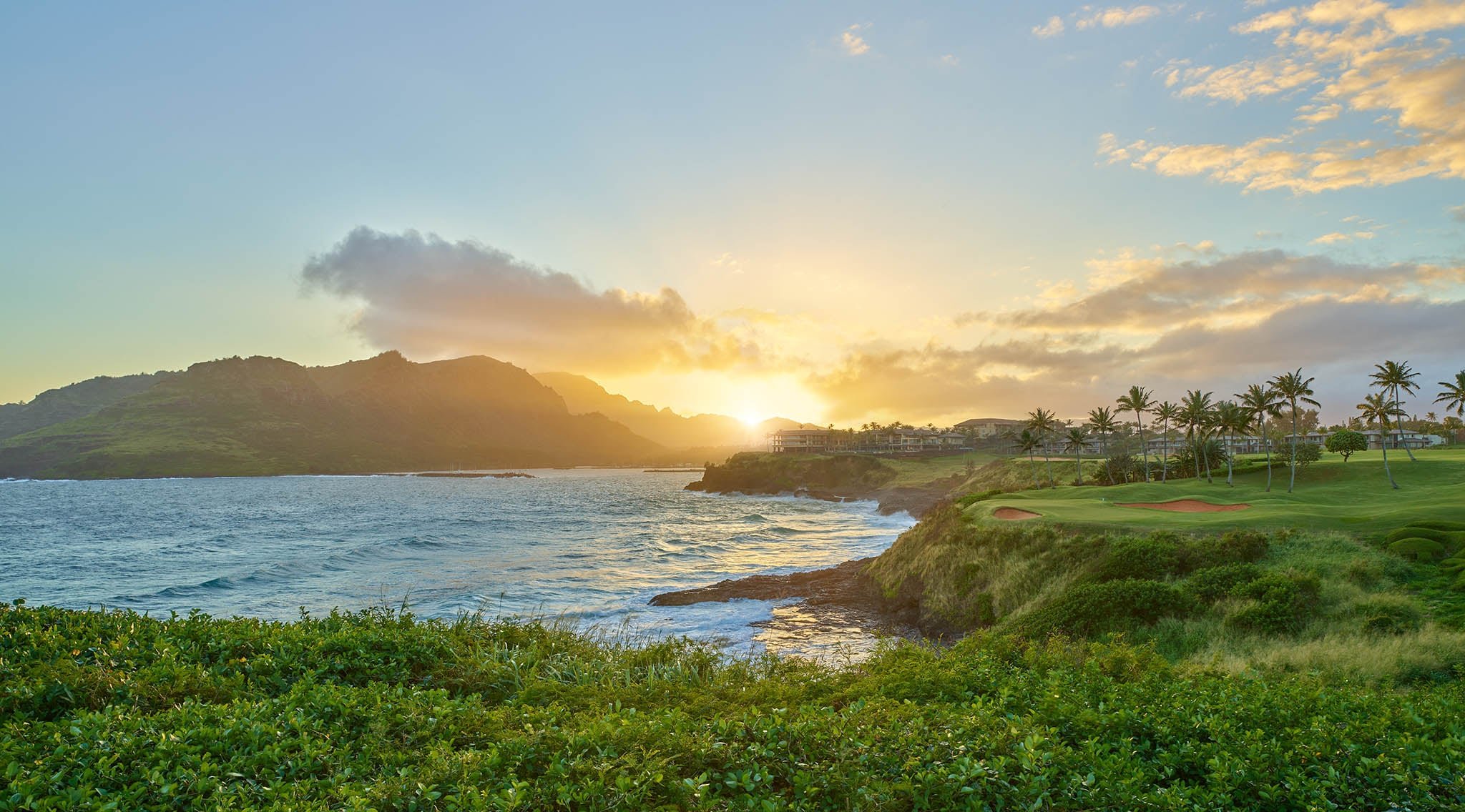 Golden hour light bathes the championship golf course as waves crash against volcanic cliffs, with swaying palms framing this dramatic oceanfront setting.