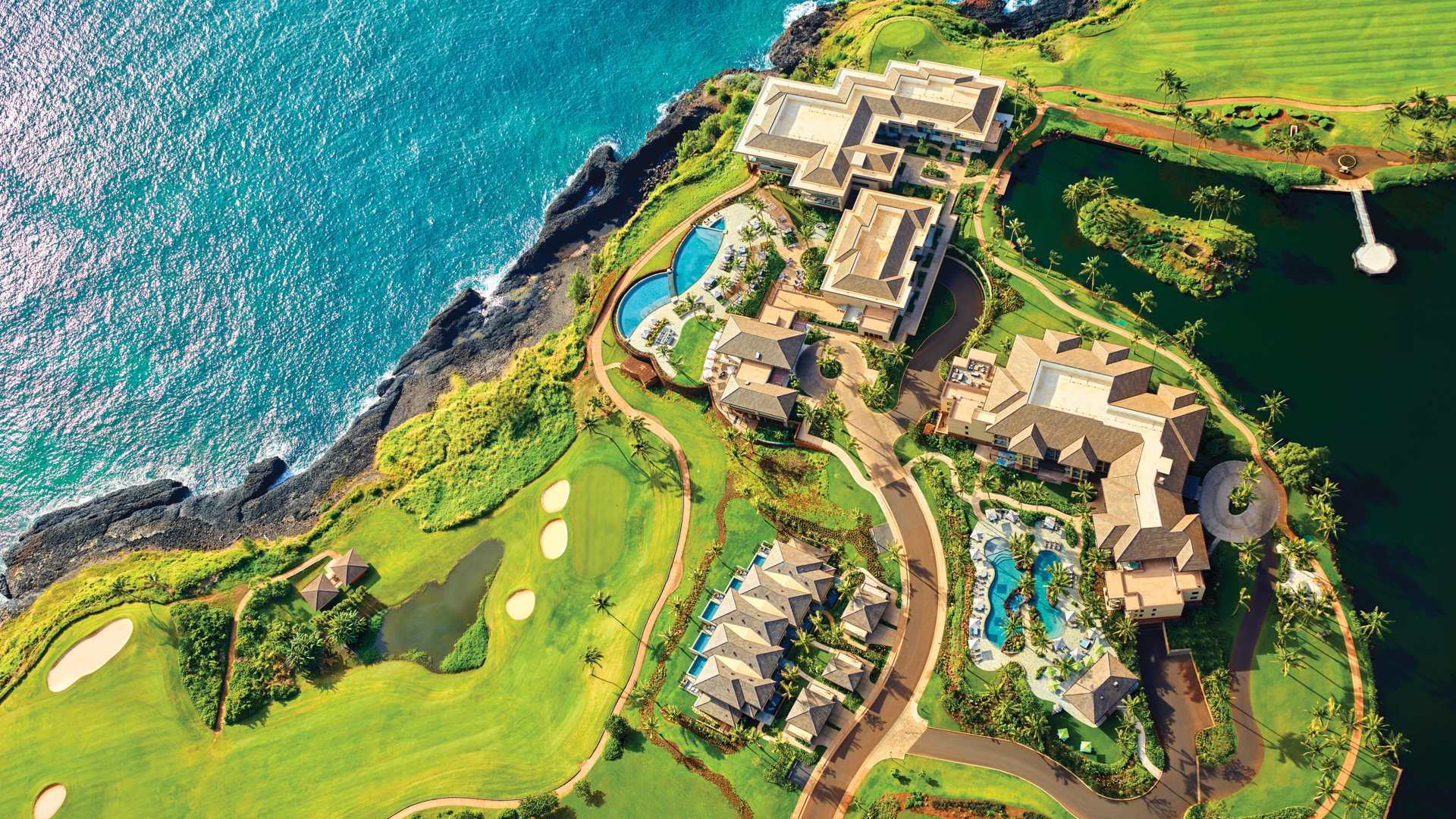 Dramatic clifftop villas cascade down emerald bluffs toward turquoise Pacific waters, while championship golf greens and serpentine pools create an island paradise above the crashing surf.