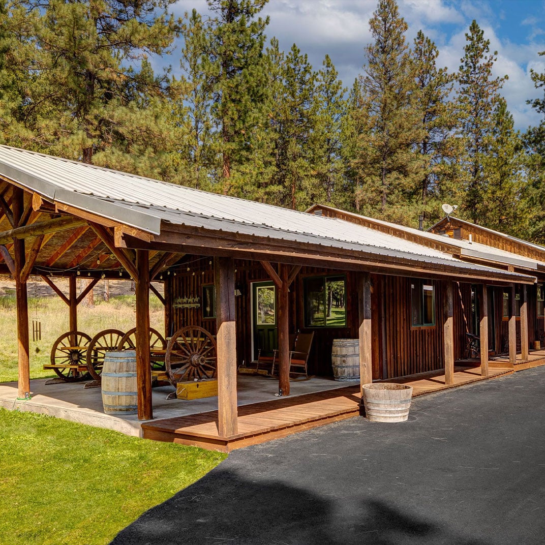 Rustic wooden buildings nestled among towering pines welcome guests with covered porches, vintage wagon wheels, and weathered barrels that speak to authentic ranch heritage.
