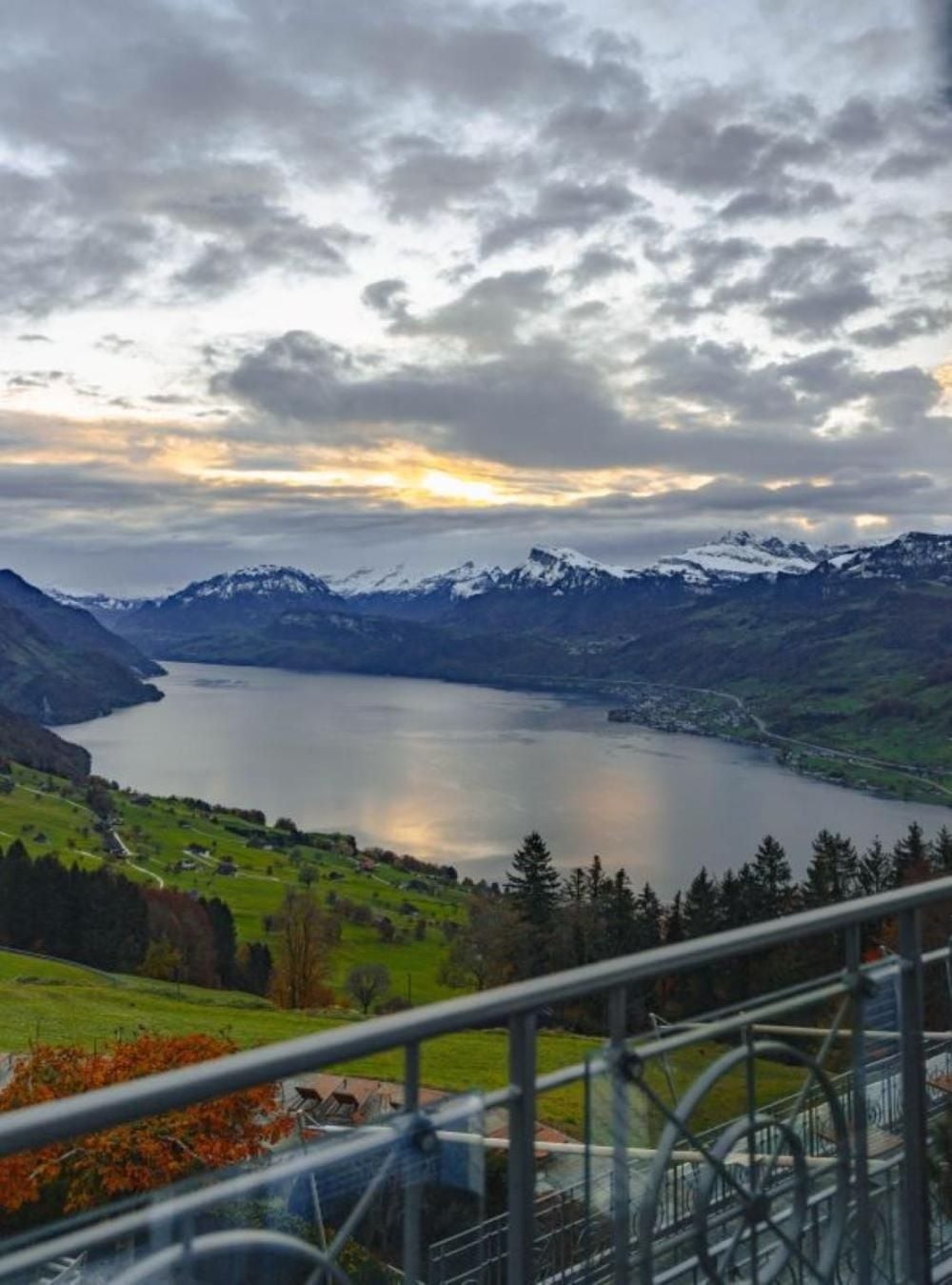 From the observation deck's steel railings, Lake Lucerne stretches like molten silver beneath snow-dusted Alpine peaks as evening light pierces dramatic storm clouds.