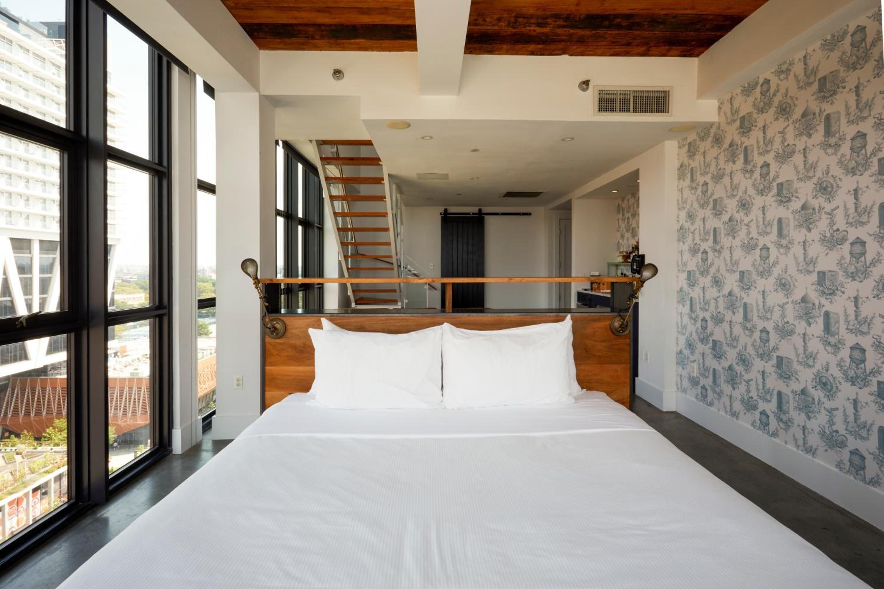 Exposed wood beams and nautical-inspired headboards create maritime charm in this loft-style suite, where industrial windows frame Brooklyn's evolving streetscape below.