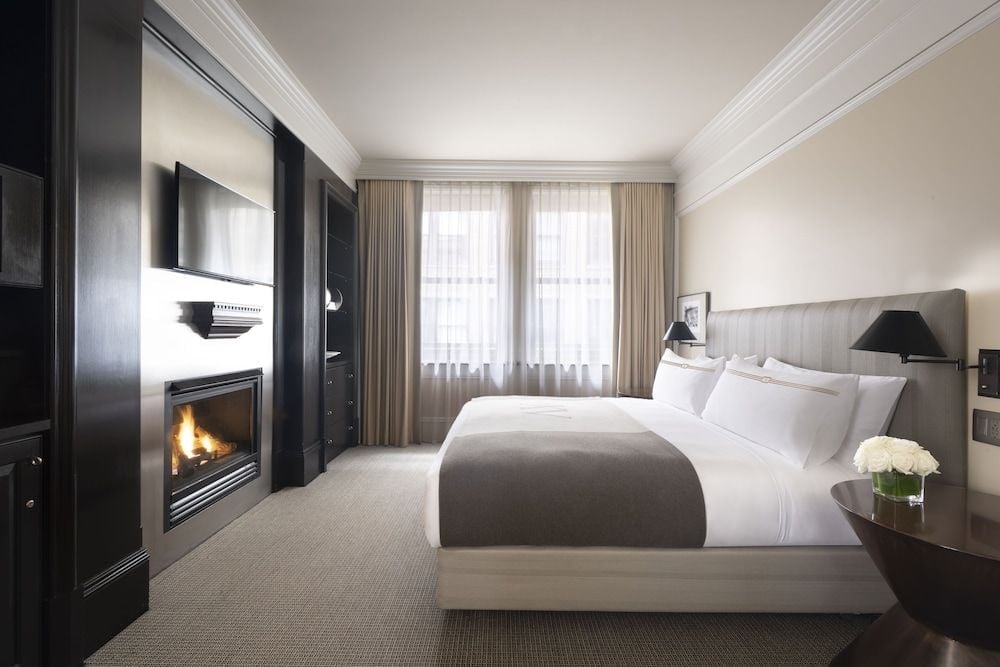 Crisp white linens contrast beautifully with rich dark millwork, while a crackling fireplace casts warm shadows across plush carpeting in this sophisticated guest room.