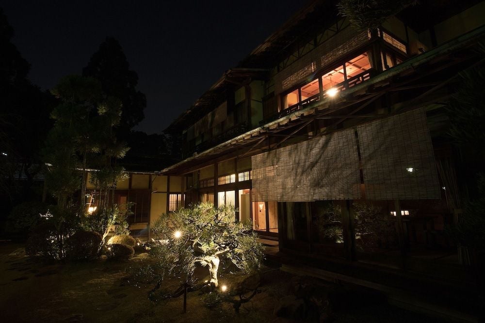 Warm amber light spills from traditional wooden windows into the meticulously landscaped courtyard, where ancient stones and carefully pruned trees create an intimate sanctuary.