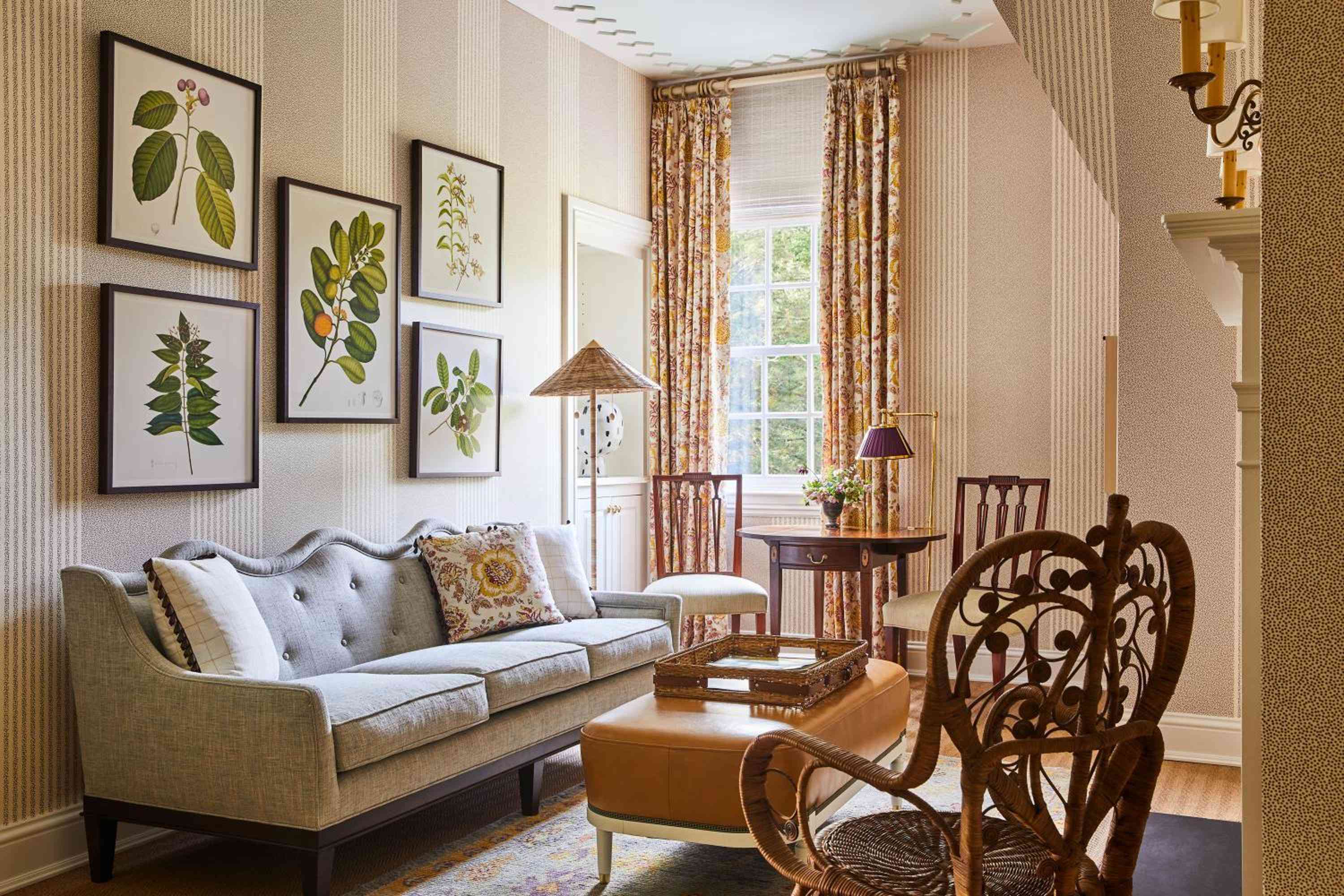 Botanical prints and vintage brass sconces frame a cozy seating area where striped wallpaper meets floral curtains in perfectly orchestrated colonial elegance.