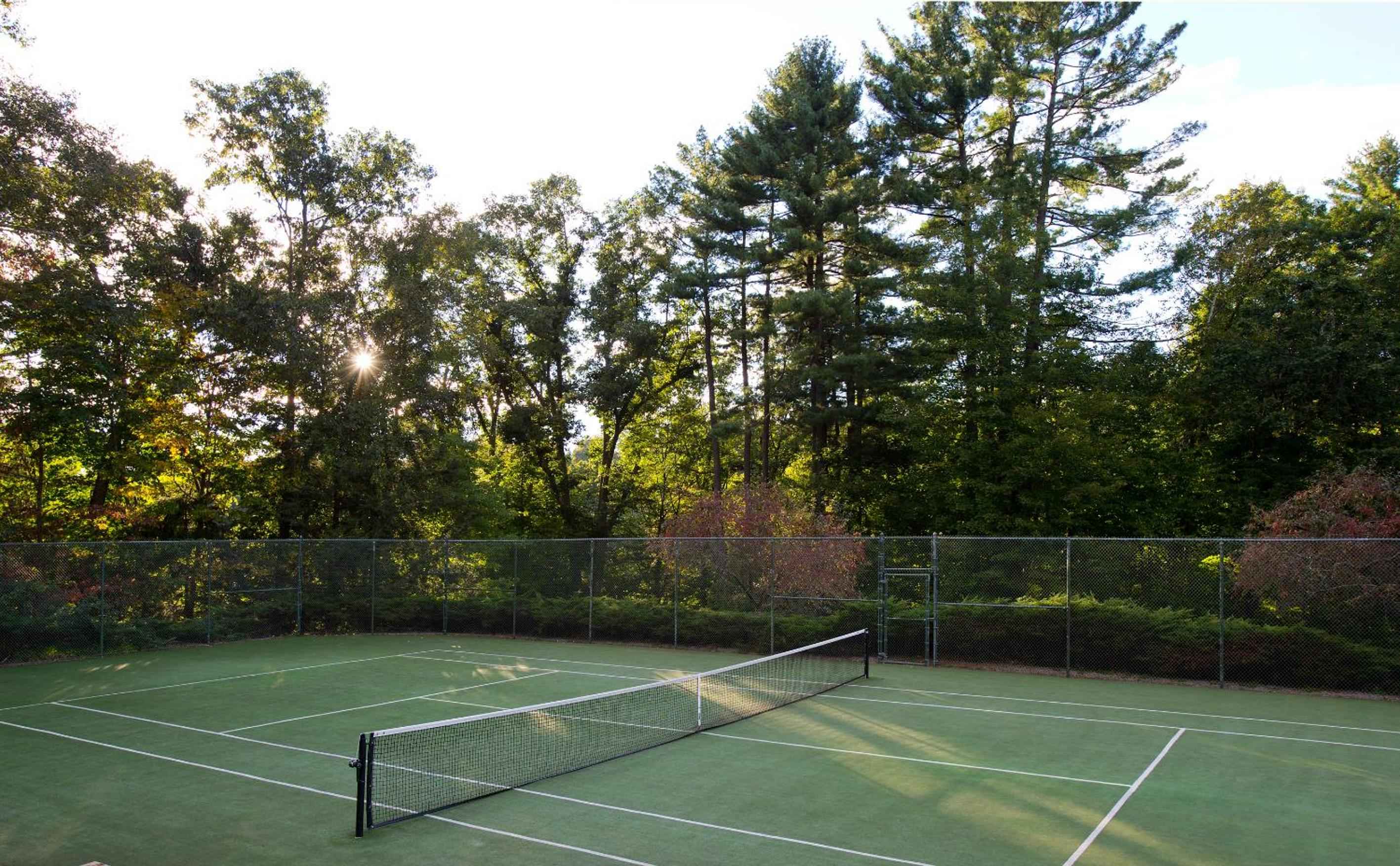 Dappled sunlight filters through towering pines onto the pristine tennis court, where afternoon shadows dance across emerald-green surfaces surrounded by lush New England foliage.