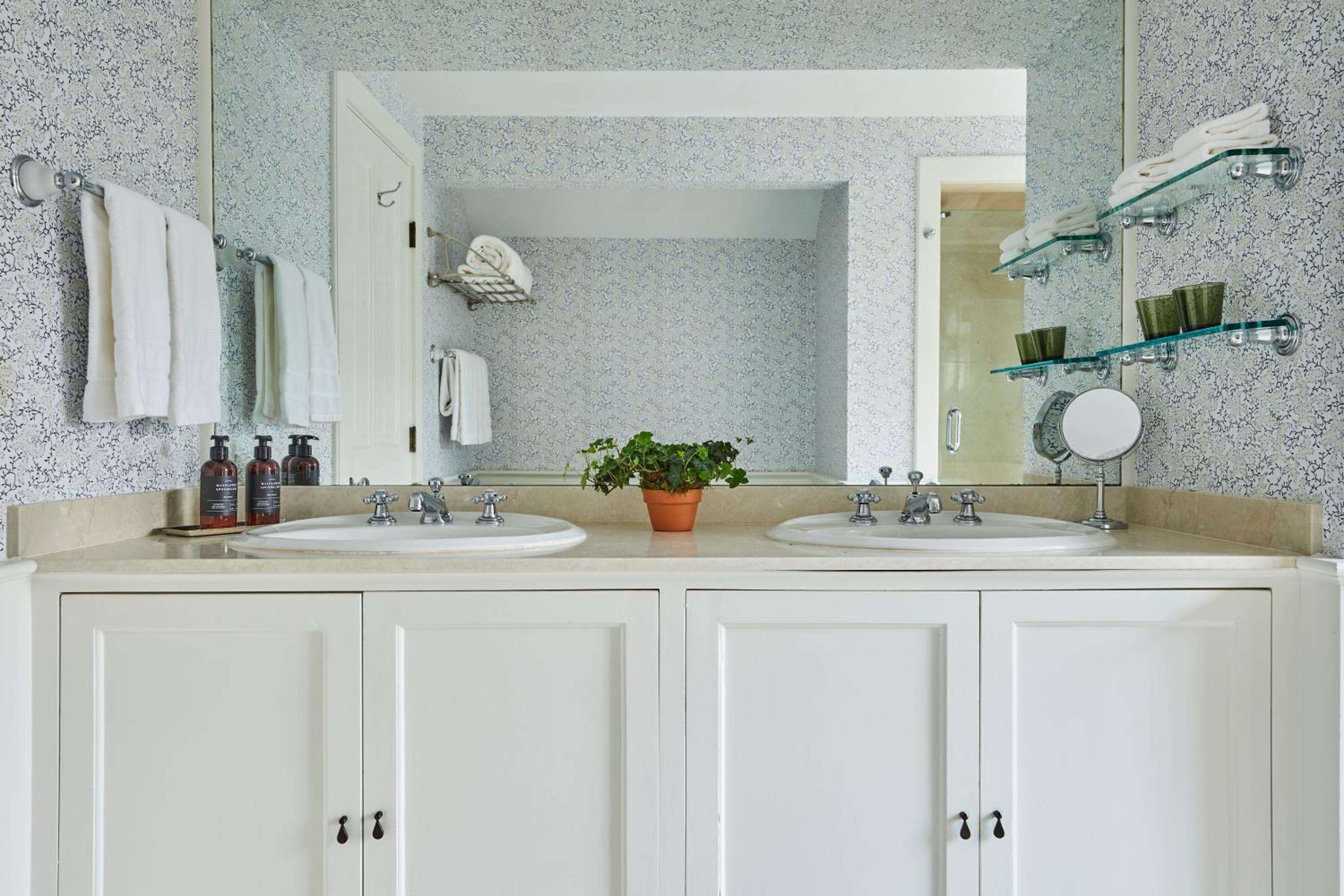 Dual vanities beneath speckled wallpaper create an intimate morning ritual space, where chrome fixtures and glass shelves reflect thoughtful design restraint.