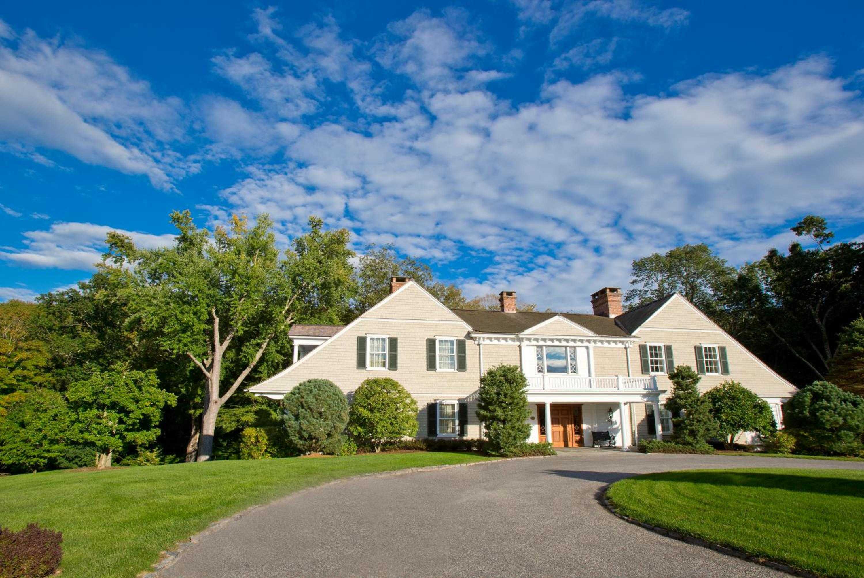 Shingle-style elegance unfolds along a graceful circular drive, where manicured lawns and towering trees frame this quintessential New England retreat's welcoming facade.