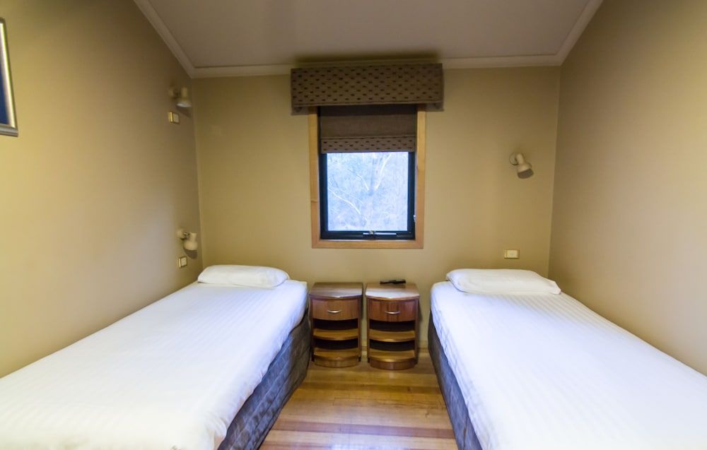 Twin beds frame a forest view through timber-trimmed windows, where natural textures and warm wood create an intimate retreat within Tasmania's coastal wilderness.