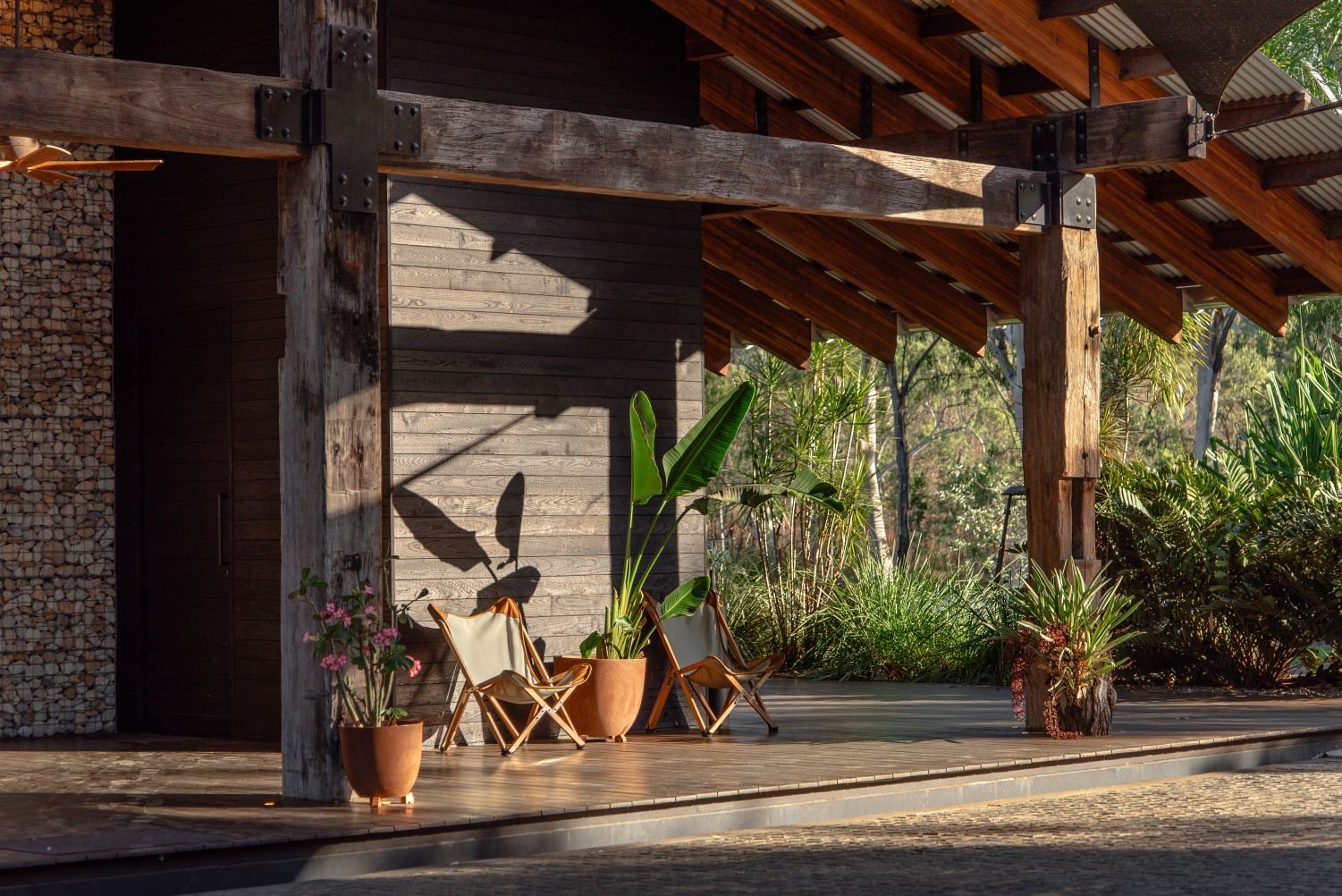 Weathered timber beams frame a serene terrace where leather safari chairs catch golden afternoon light, while tropical plants soften the lodge's rugged industrial architecture.