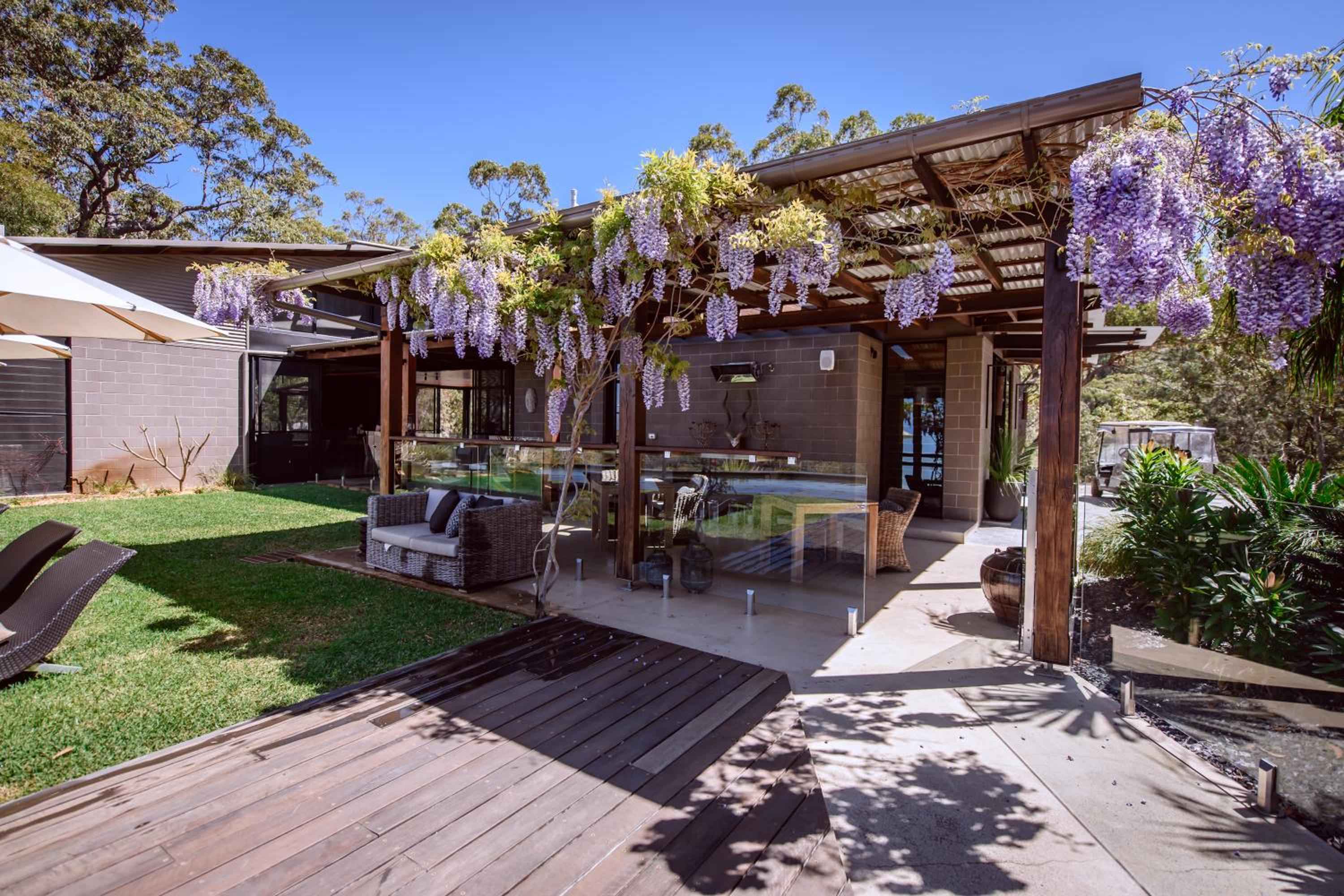 Purple wisteria cascades over timber pergolas, creating natural canopies above contemporary outdoor lounging areas where glass balustrades frame manicured lawns and native eucalyptus beyond.