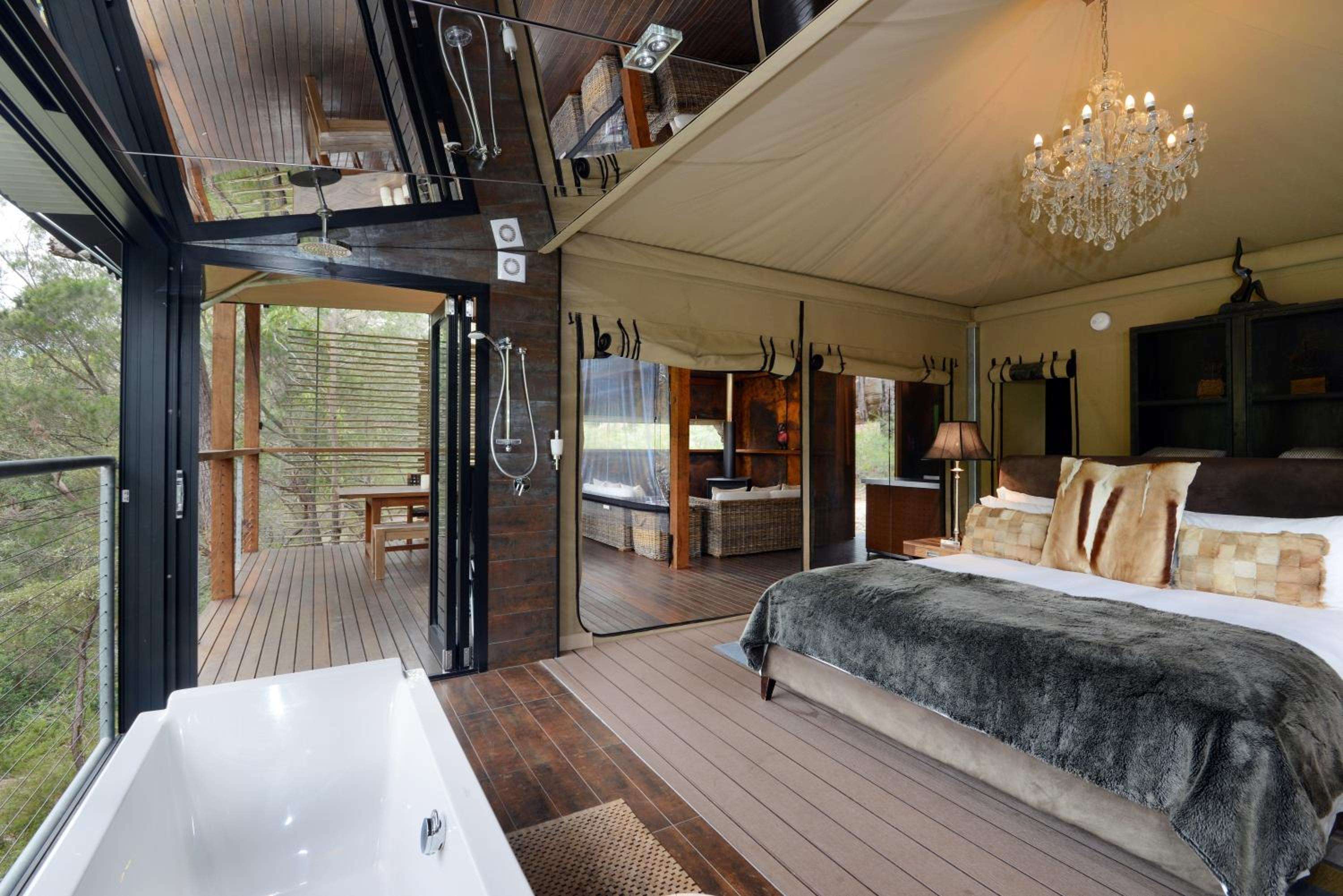 Crystalline chandelier light dances across polished timber floors where freestanding tub meets king bed, while floor-to-ceiling glass dissolves boundaries between intimate luxury and untamed bushland beyond.