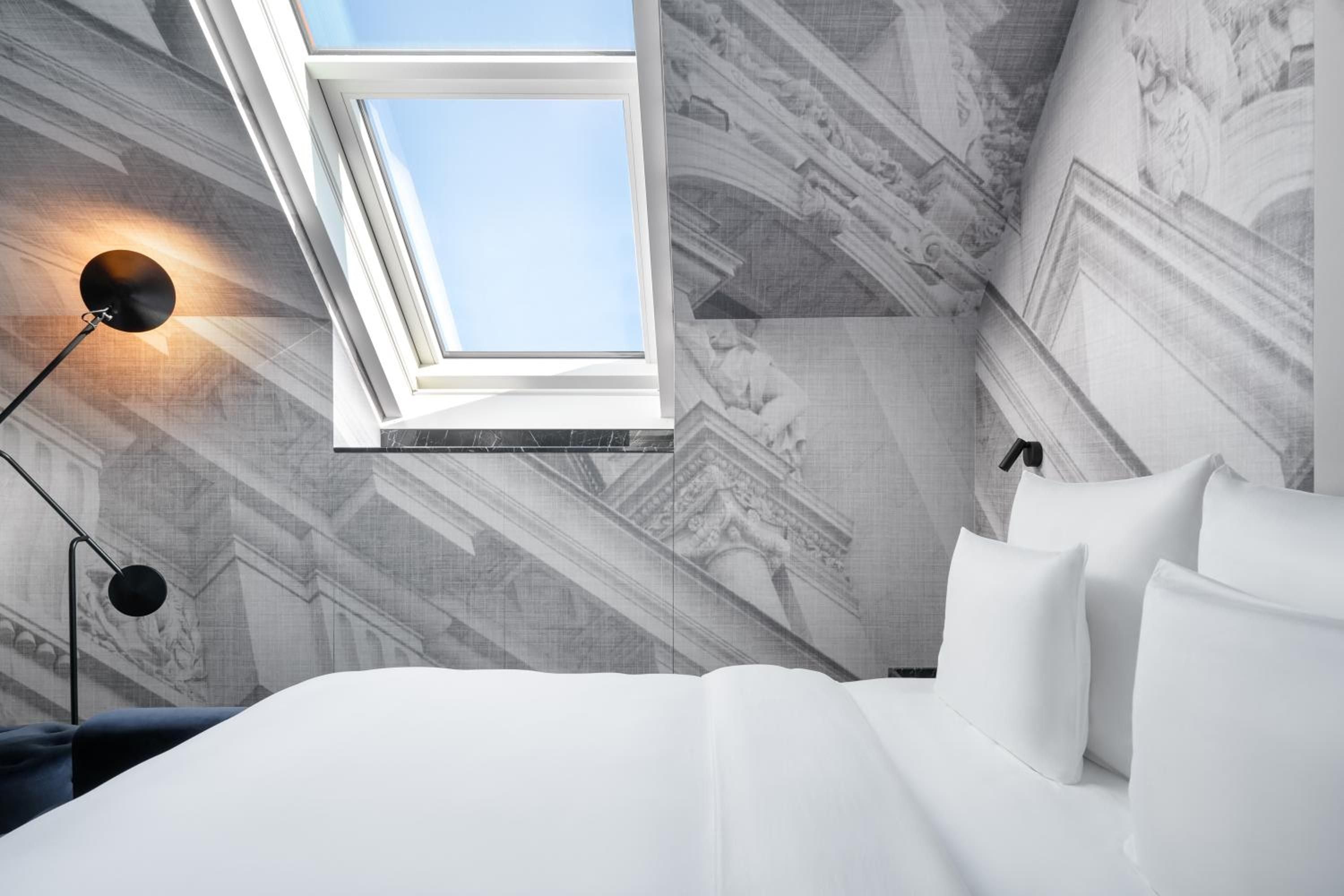 Skylights frame blue Vienna sky above this intimate attic retreat, where classical architectural prints create an unexpected gallery wall beneath exposed beams and contemporary black lighting.