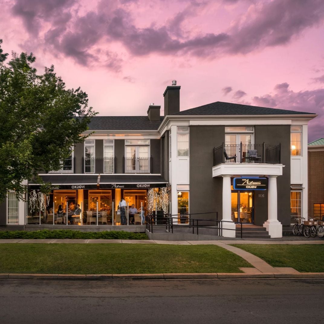 Pink-hued twilight bathes this elegant corner building where warm restaurant lighting spills onto manicured lawns, creating an inviting urban oasis with classical architectural details.