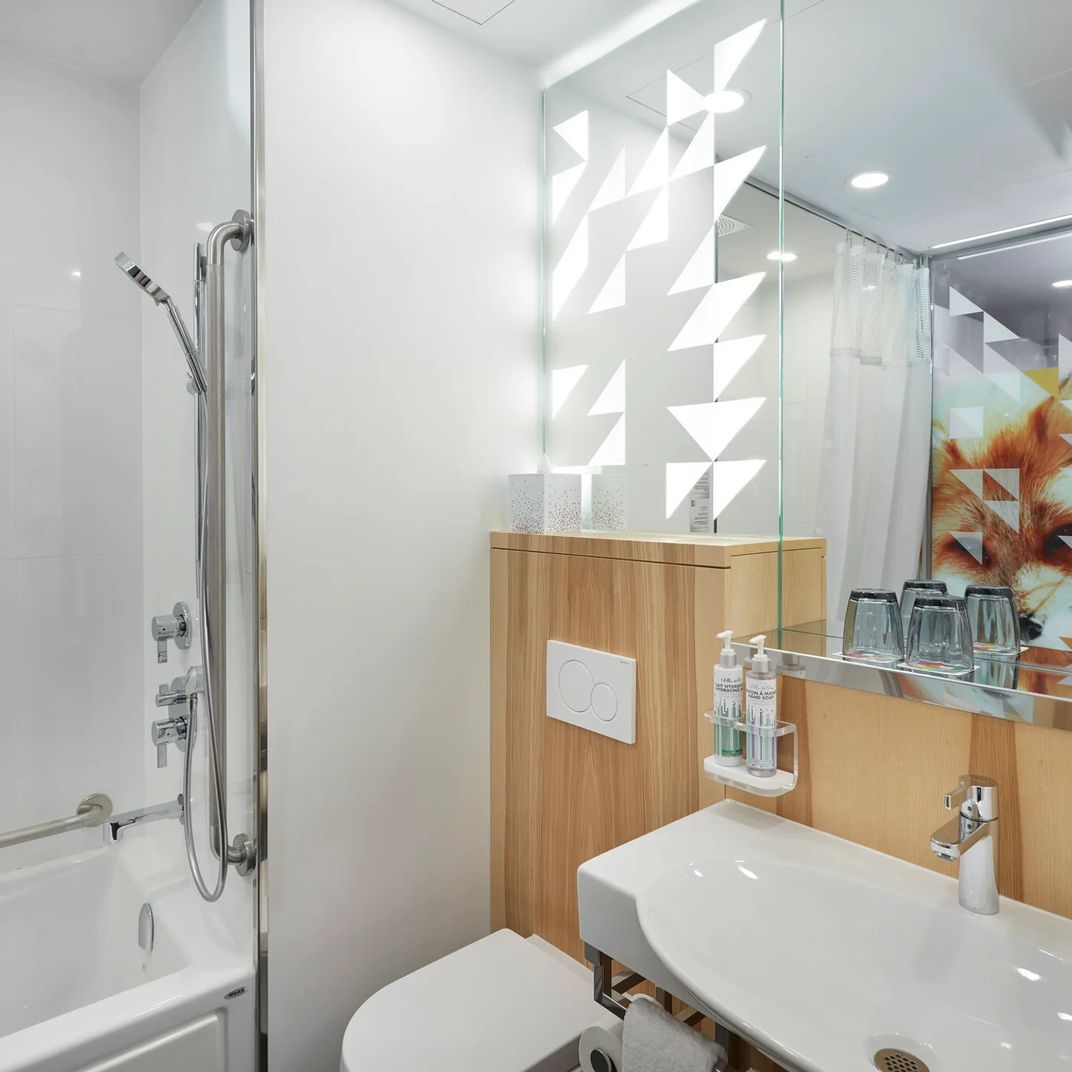 Geometric shadows dance across pristine white walls while warm wood vanity and glass-enclosed shower create an intimate sanctuary of Scandinavian-inspired calm.