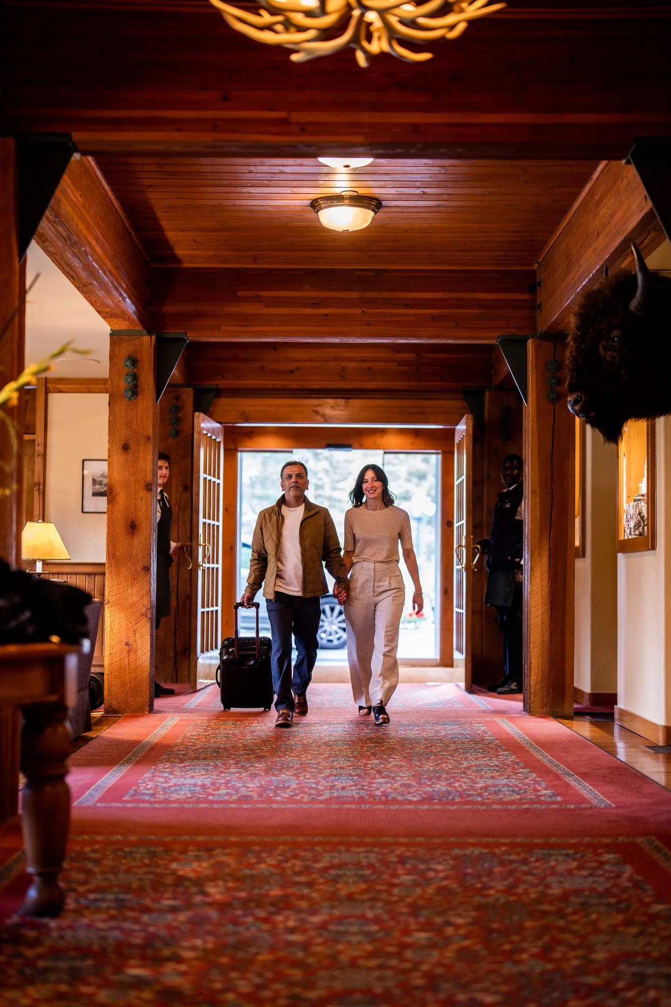 Couples stroll through the Post's timber-framed lobby, where exposed beams and Persian runners create an intimate mountain lodge atmosphere beneath warm pendant lighting.
