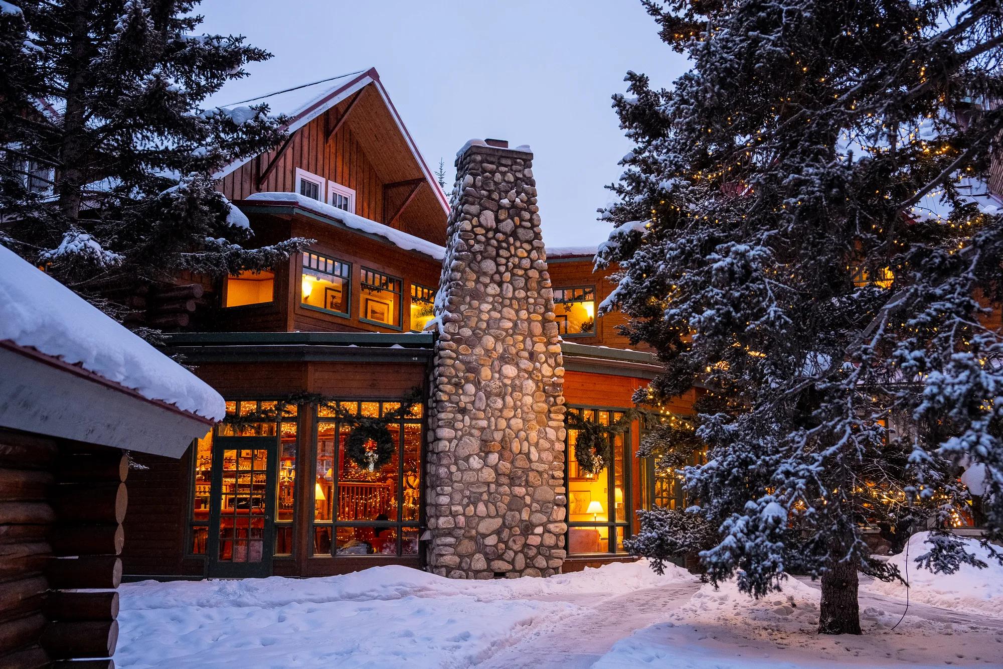 Snow-dusted evergreens frame the lodge's glowing windows and towering stone chimney, where warm amber light spills onto pristine powder in the mountain twilight.