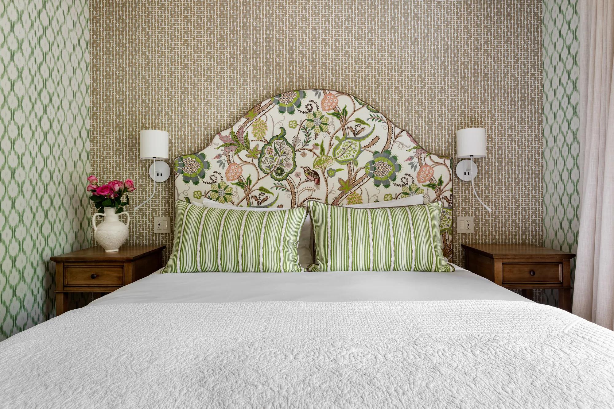 Floral upholstered headboard and green striped pillows create cottage charm against grasscloth wallpaper, while warm sconces cast intimate light over crisp white linens.